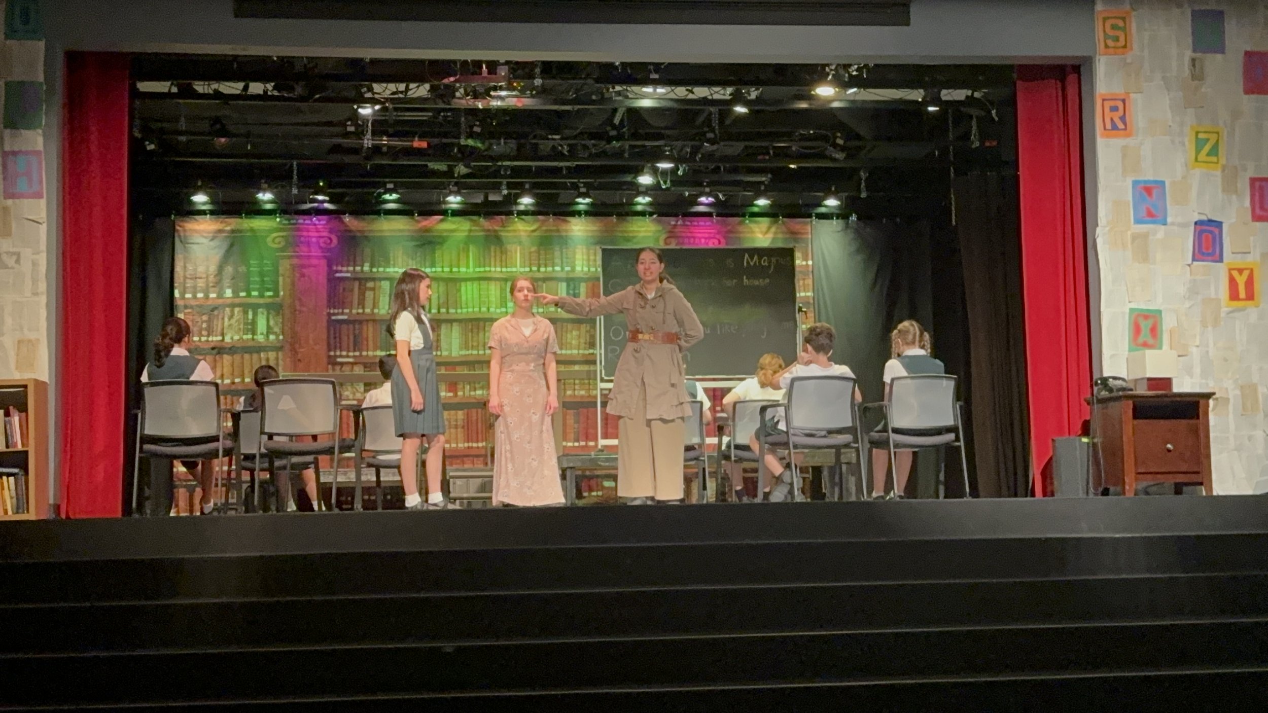 School play on stage depicting a classroom scene with students and teachers, set with bookshelves and chalkboard in the background, and red curtains on either side.
