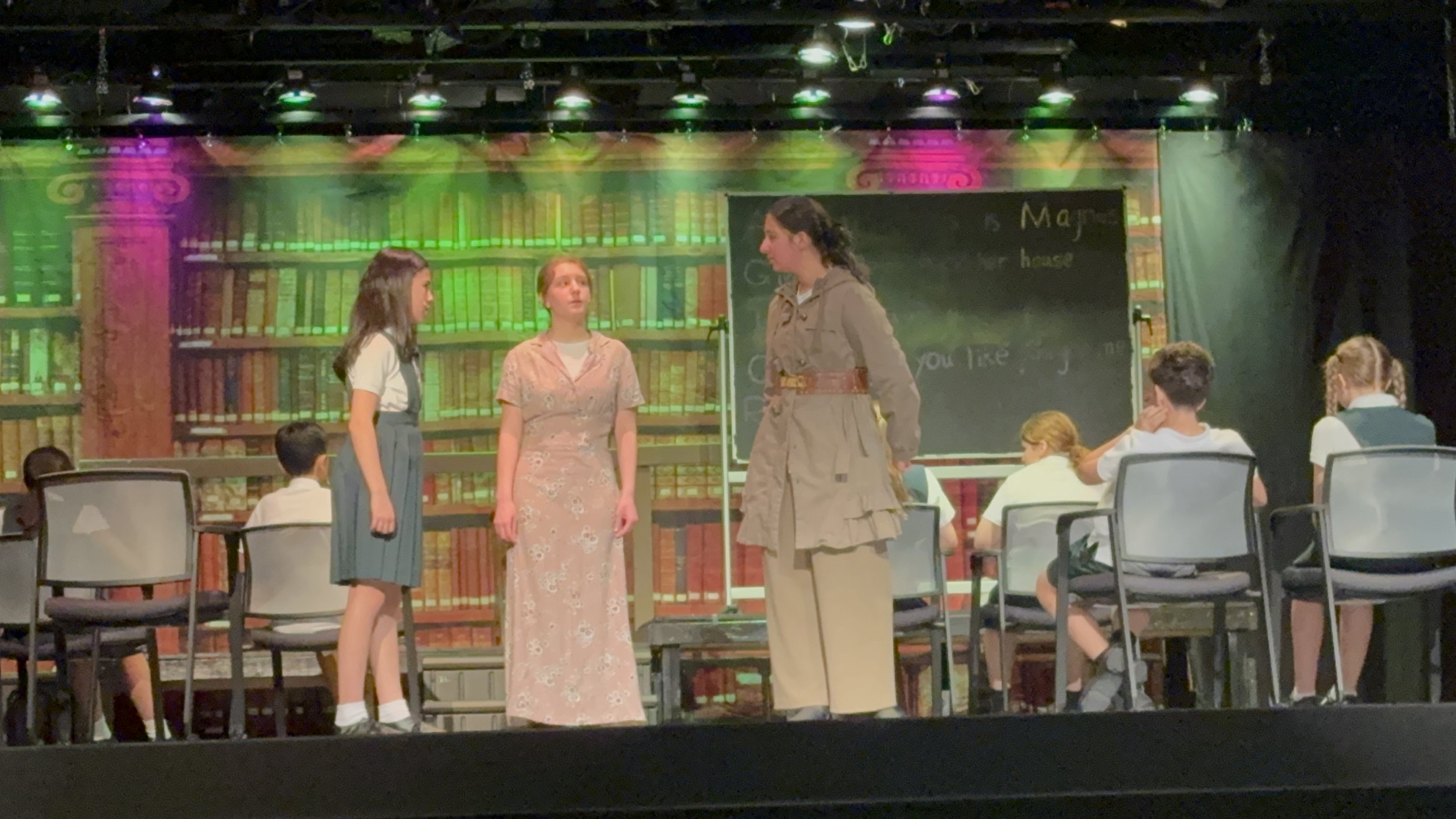 A stage scene from a school play with three girls standing and five students seated, set in a library background with bookshelves and a blackboard.