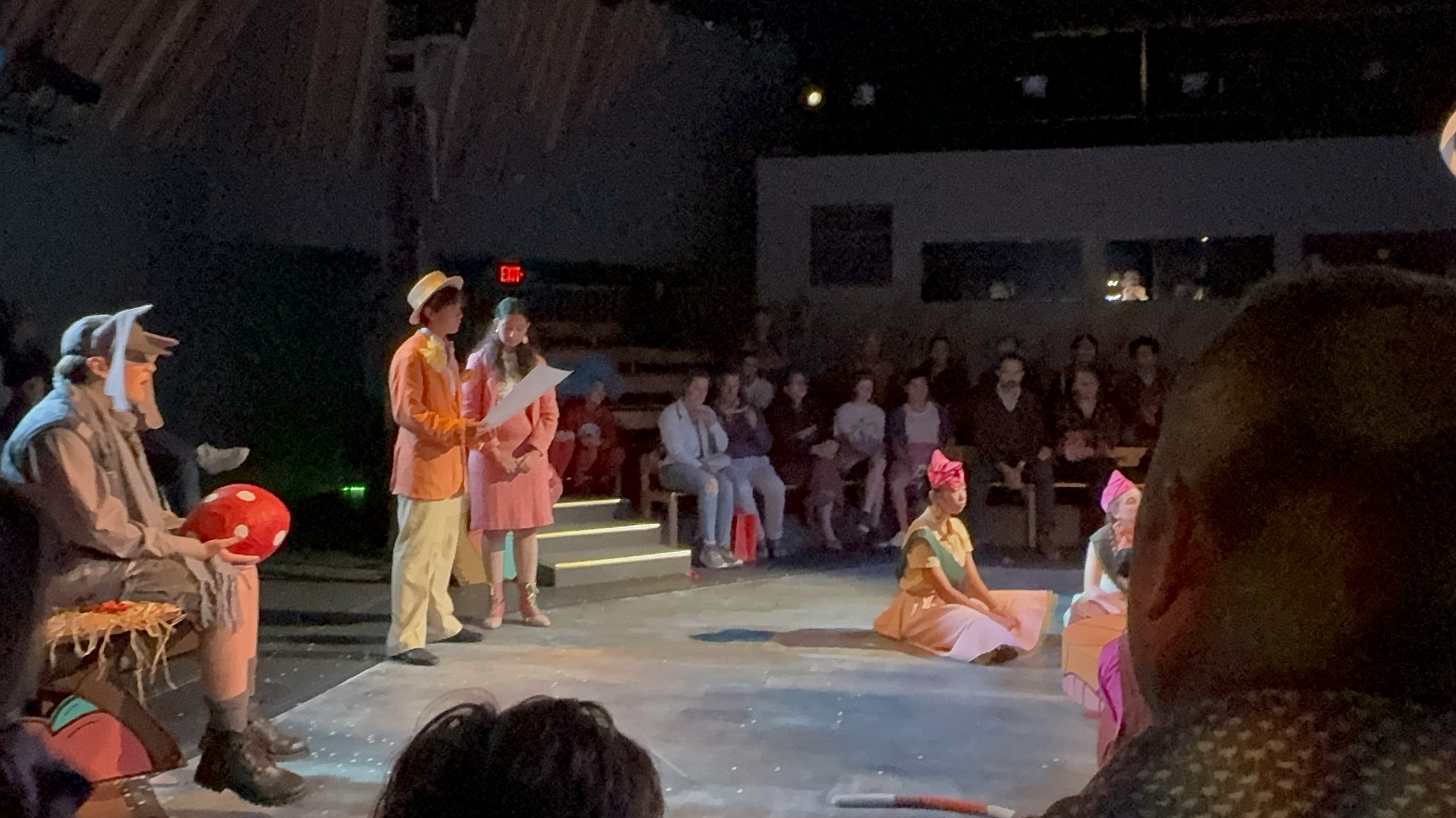 A theatrical performance on stage with actors dressed in colorful costumes, some sitting on the floor and some standing, with an audience seated in the background in a theater.