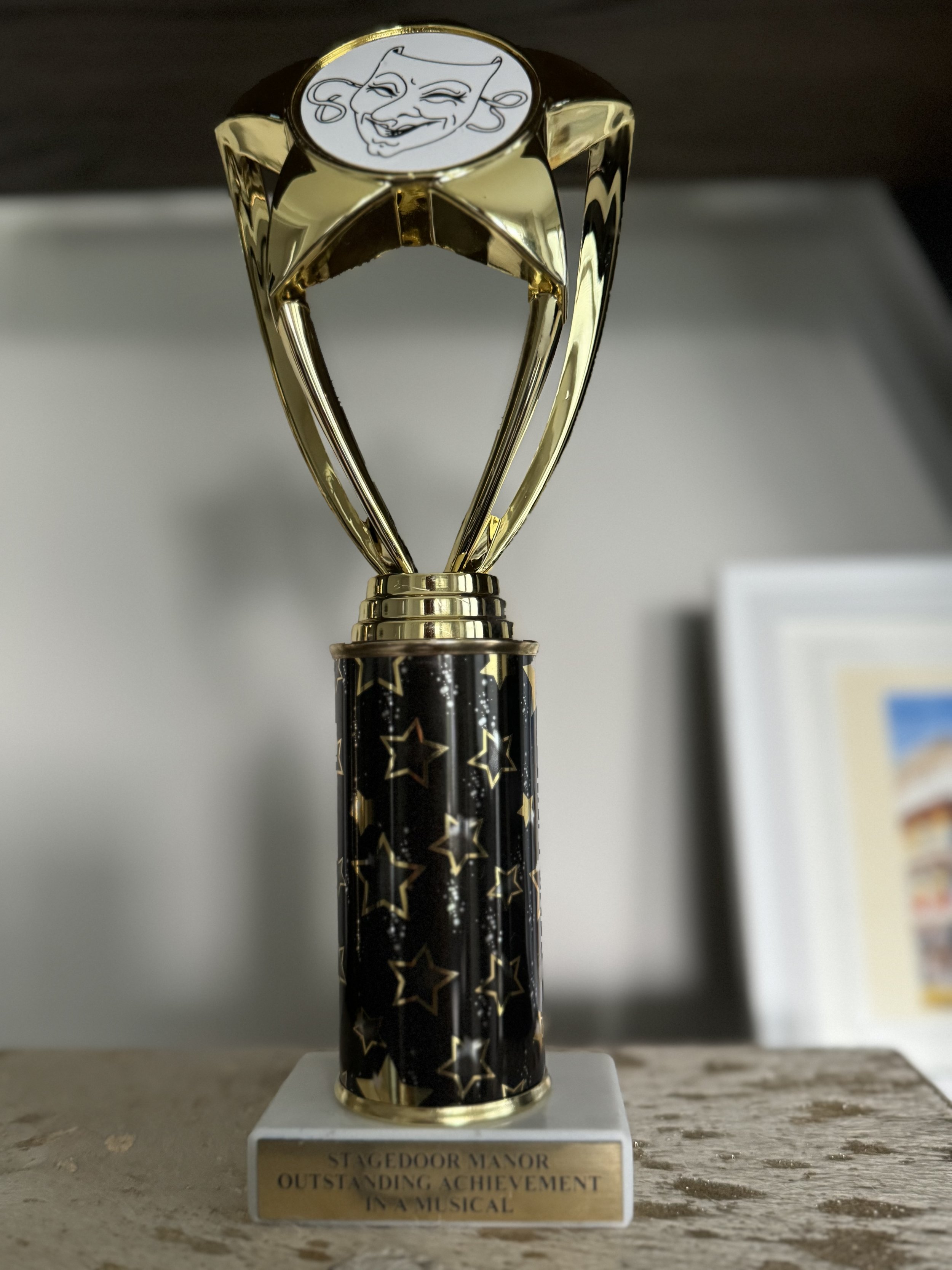 Gold trophy with a black base decorated with gold stars, featuring a cartoon face with a winking expression on top, and a plaque that reads 'Stage Door Manor Outstanding Achievement in a Musical.'