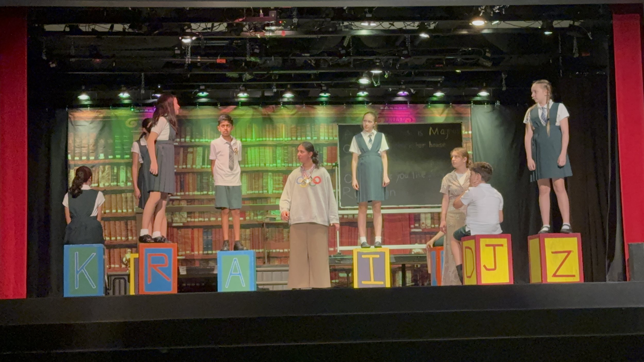 Children in school uniforms perform on stage, standing or sitting on colorful blocks with letters, in a play set in a library room.