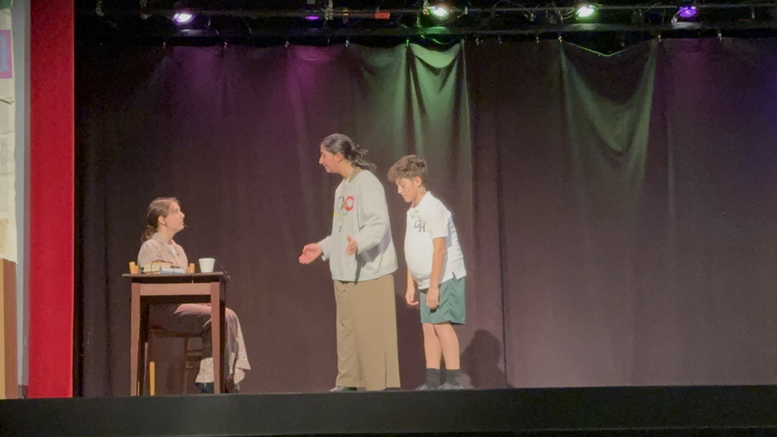 Three people on stage performing a play, one woman sitting at a table and two standing and talking, with a dark backdrop and stage lighting.