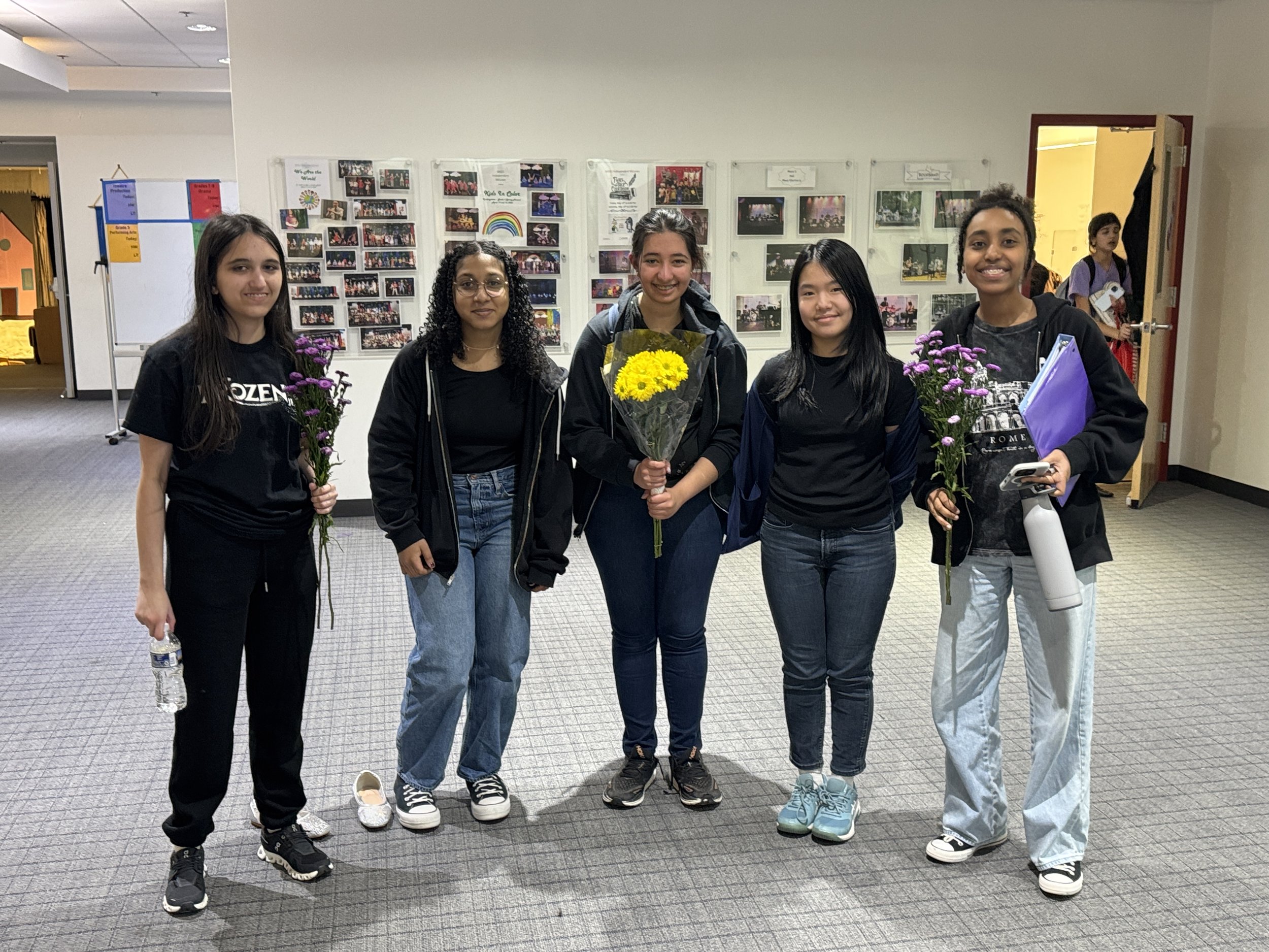 Five young women standing together in an indoor space, holding flowers and a water bottle, smiling at the camera.