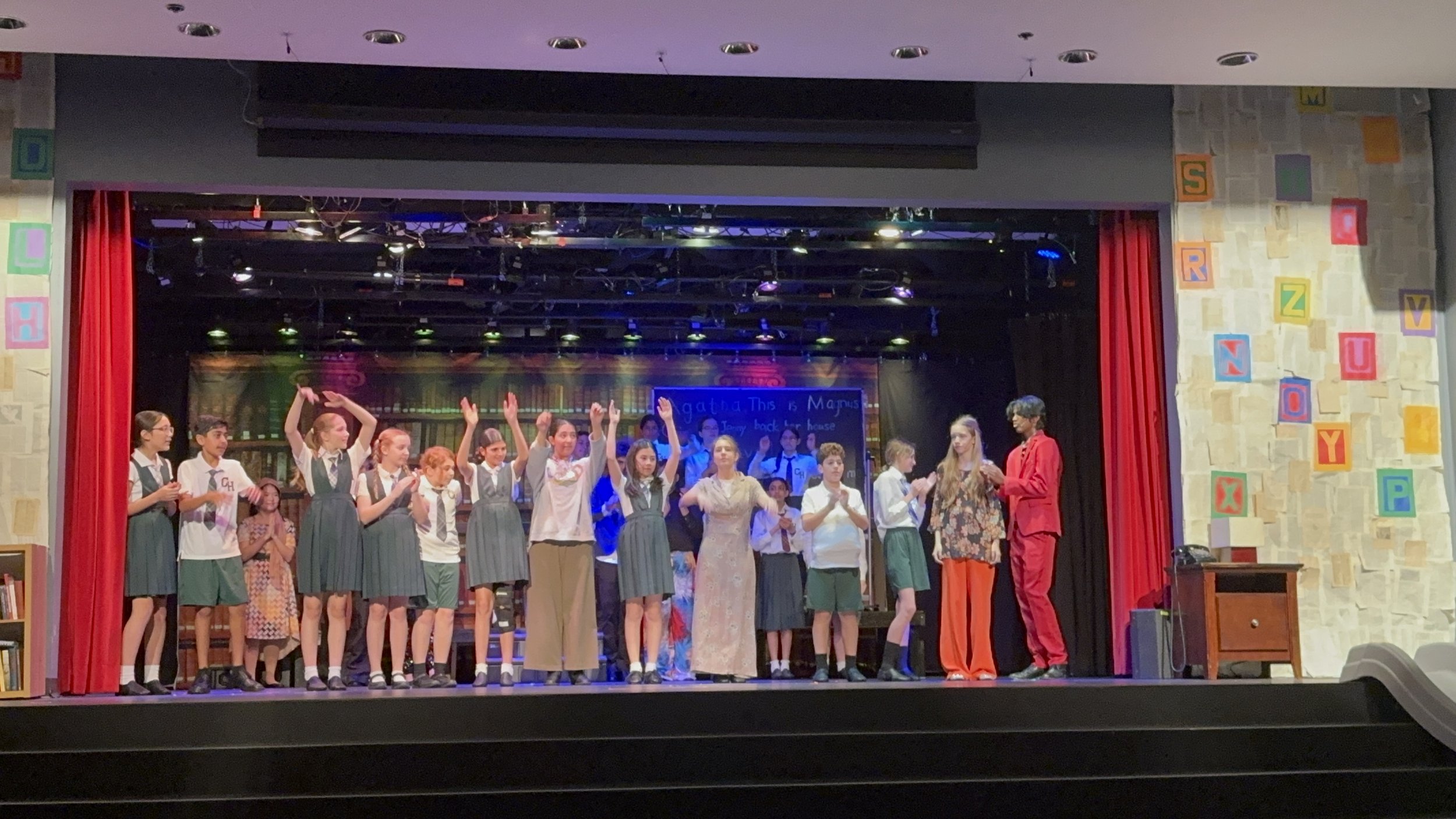 Students on stage in school play, dressed in costumes and uniforms, with some clapping and others raising their hands, and two adults standing to the right.