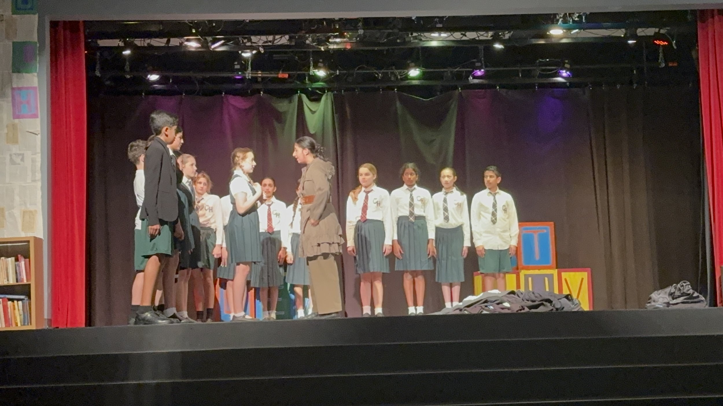 School children in uniform perform a play on a stage with a dark curtain background, colorful blocks on the right, and red curtains on the sides.
