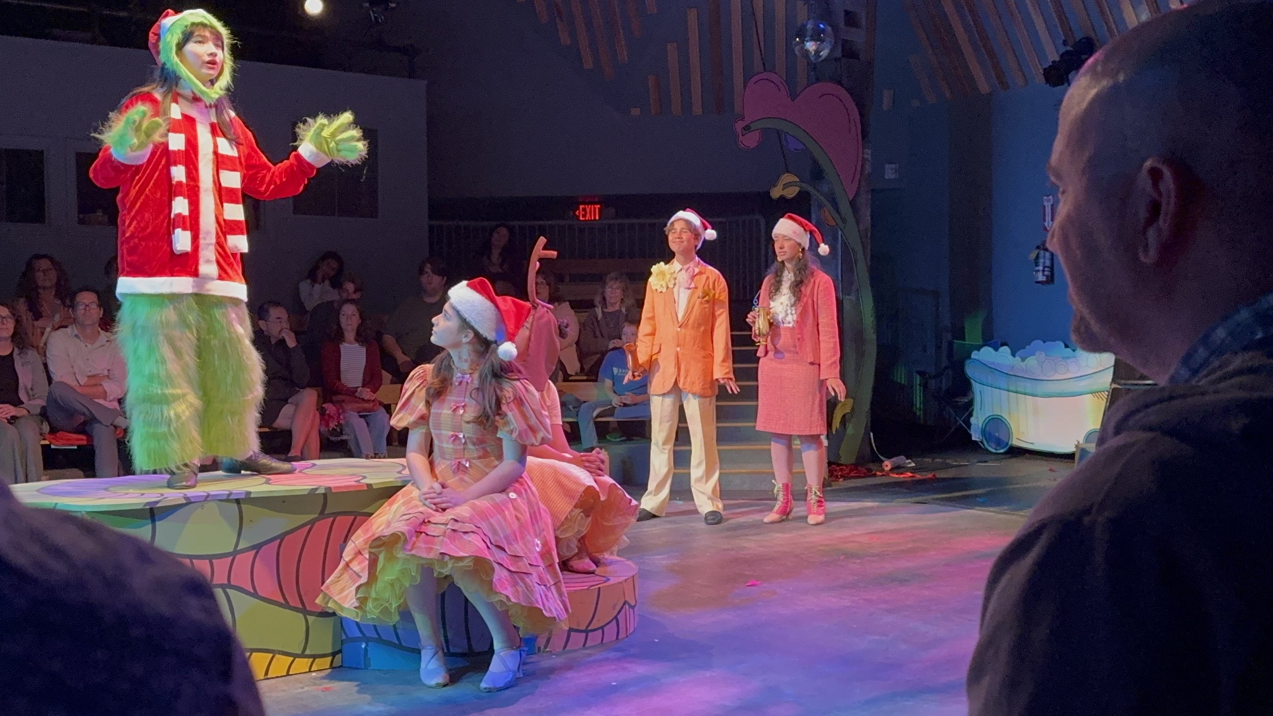 The image shows a theatrical performance with actors dressed in colorful costumes, including Christmas-themed attire. One actor is standing on a stage elevated above the seated audience, while others are sitting or standing nearby. The stage has vibr
