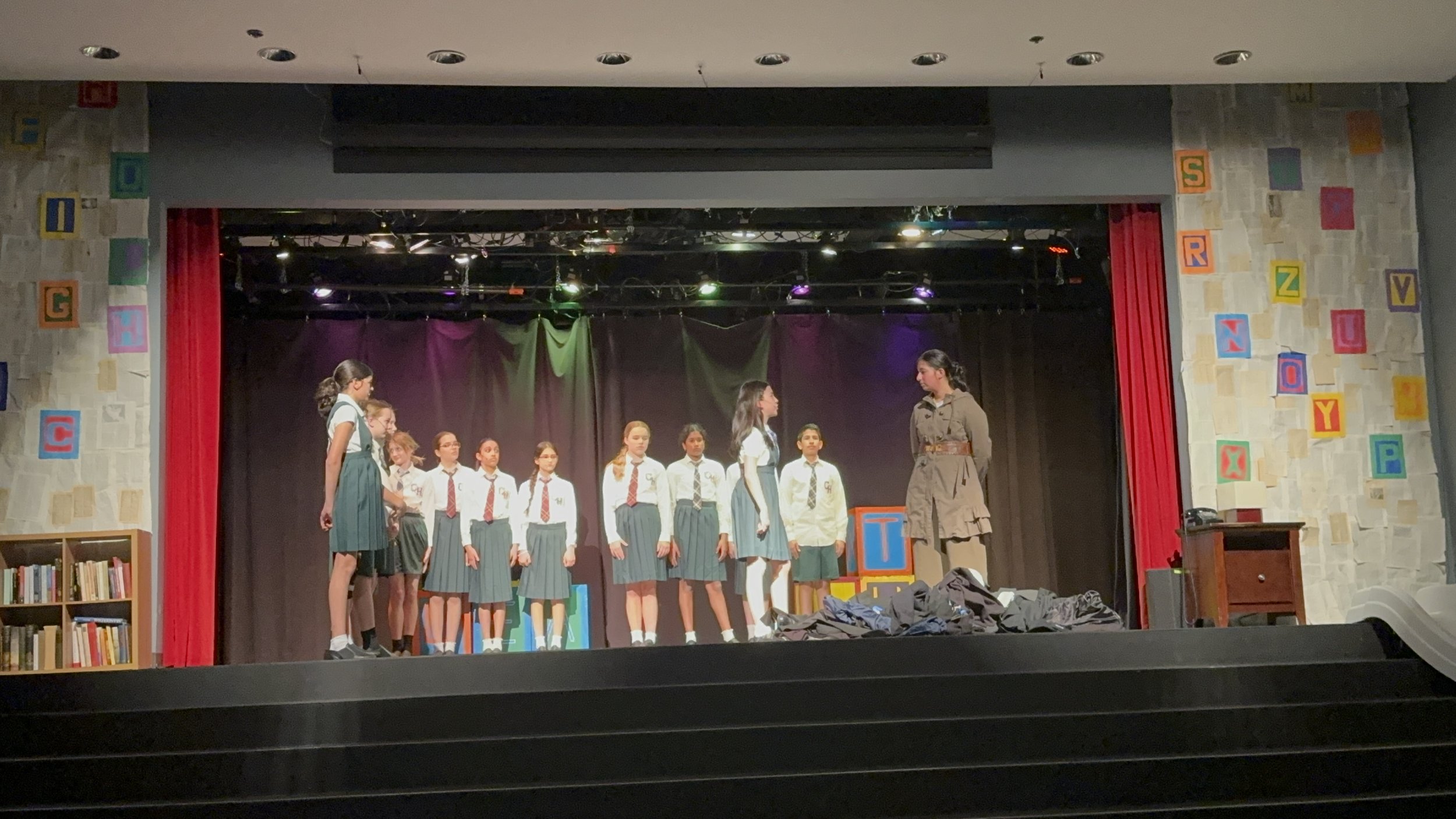 Group of children performing on a theater stage with two women at the front; one woman is speaking while the children stand in two rows behind her; colorful alphabet blocks decorate the stage area.
