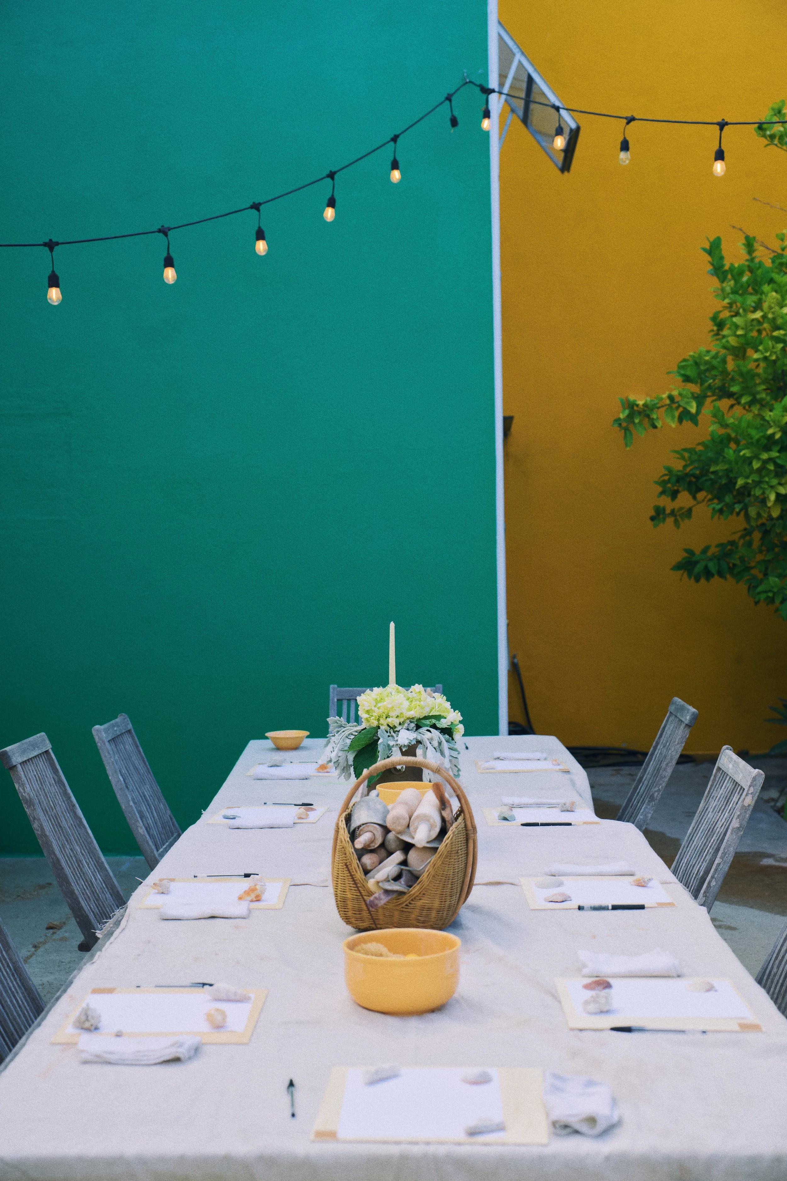 Outdoor dining table setup with placemats, napkins, and a basket of rolled-up blankets or towels. A large floral centerpiece and small bowls are also on the table. In the background, there are green and yellow walls and string lights hanging above.