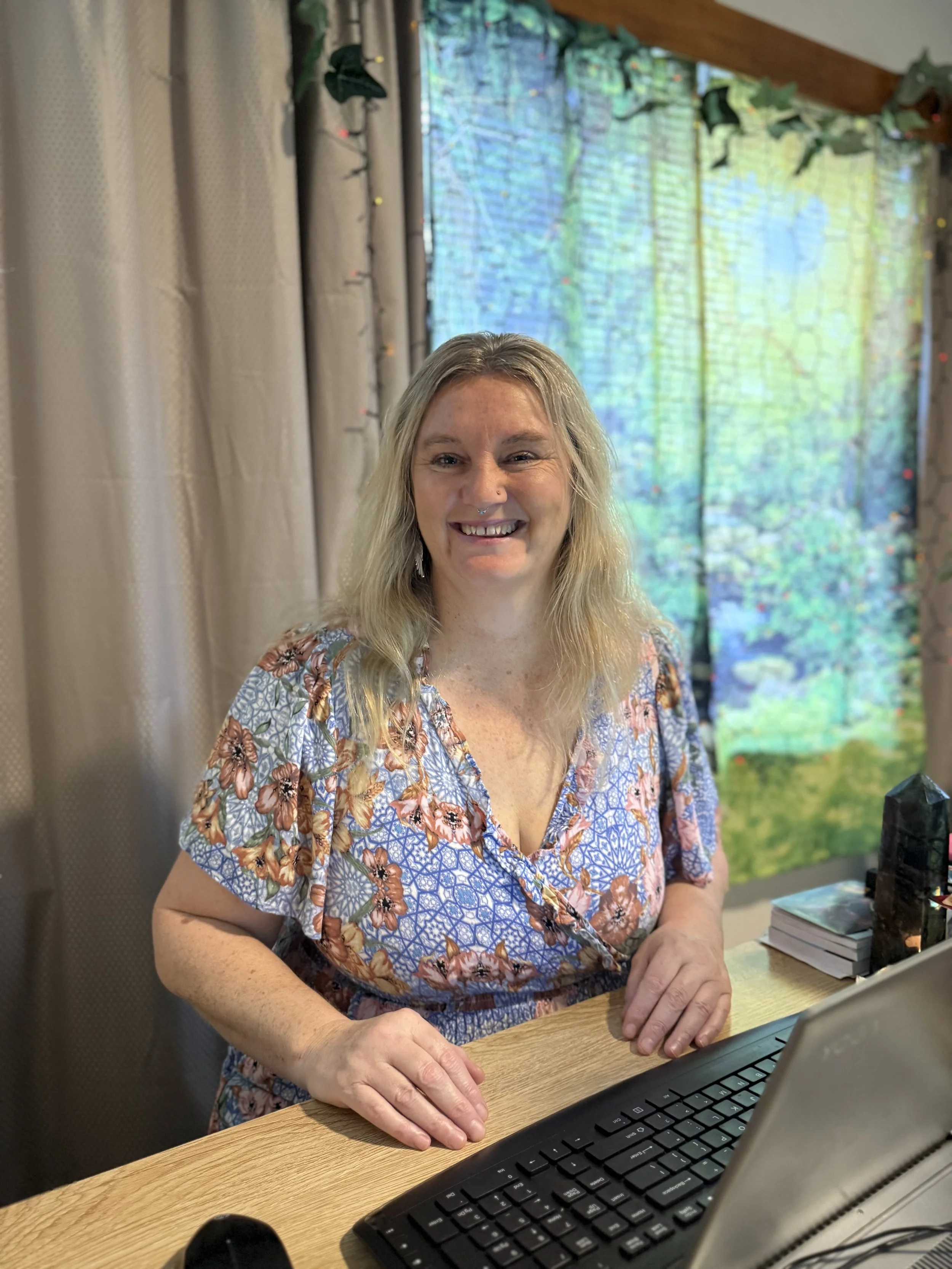 Mystic Nic wearing a floral dress, sitting at a desk with a keyboard, smiling at the camera. Behind her is a window with a nature-themed curtain. A perfect image of online offerings.