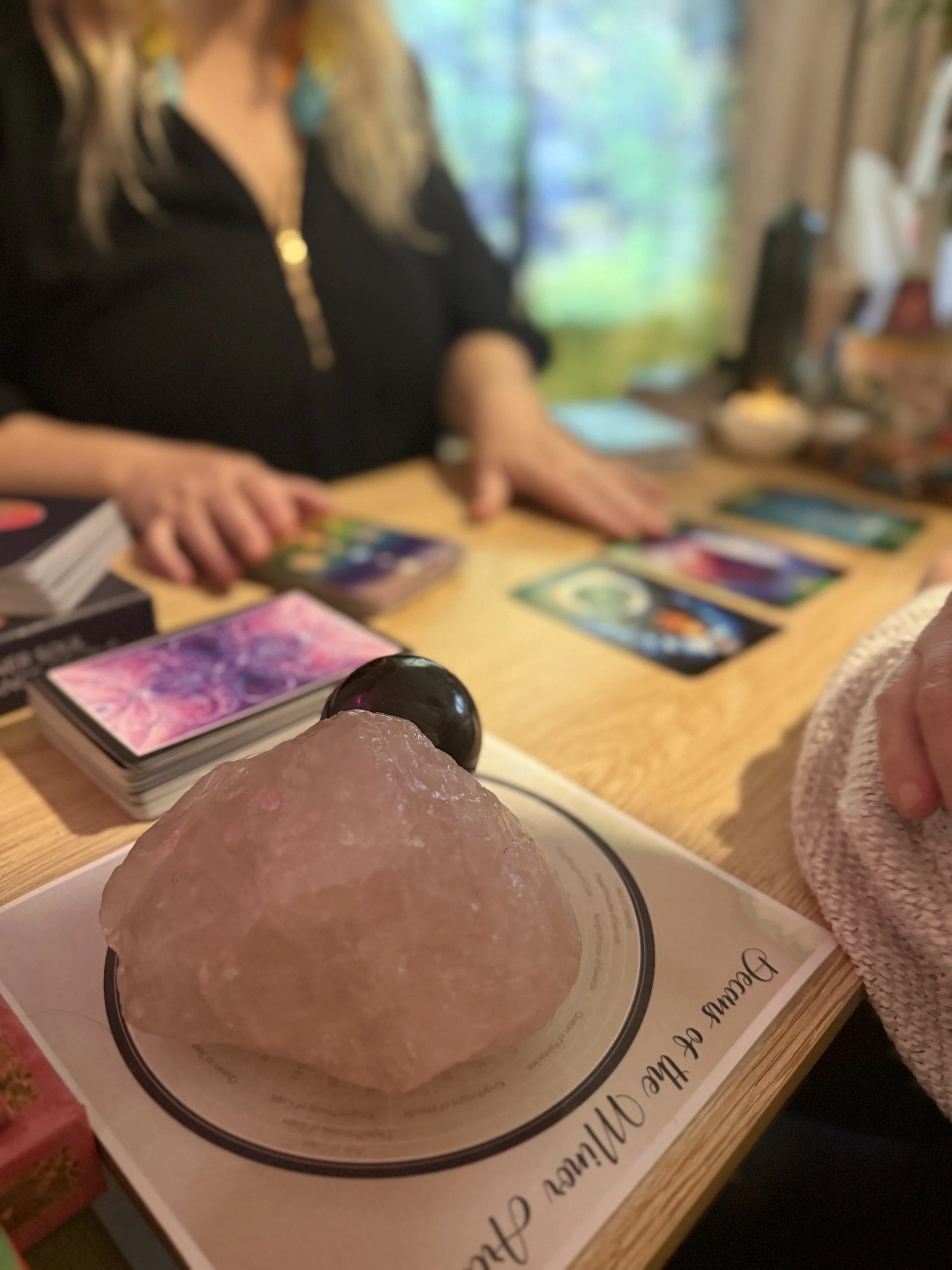 Close-up of a pink Himalayan salt lamp with a black sphere on top, placed on a booklet on a wooden table. In the background, Mystic Nic is offering a reading with tarot or oracle cards.
