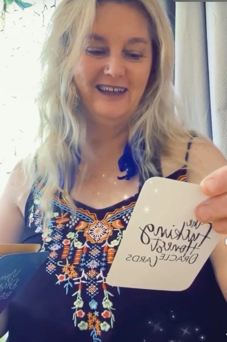 Nicole wearing a colorful embroidered dress holding a white card with writing, smiling inside a room with curtains and christmas lights