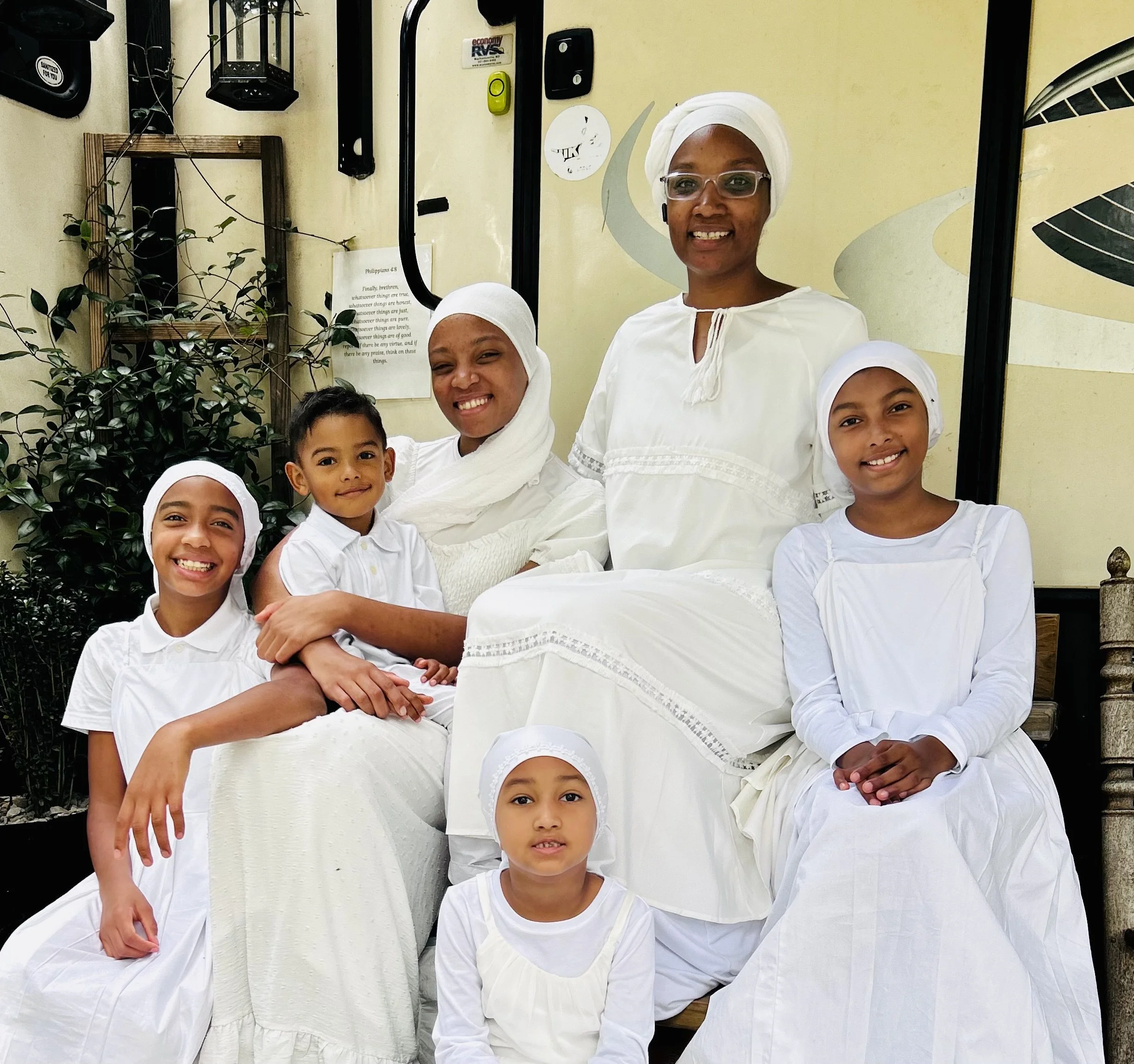 A group of seven people, including four children and three women, dressed in white, sitting and standing together in what appears to be a welcoming indoor or porch setting, smiling at the camera.