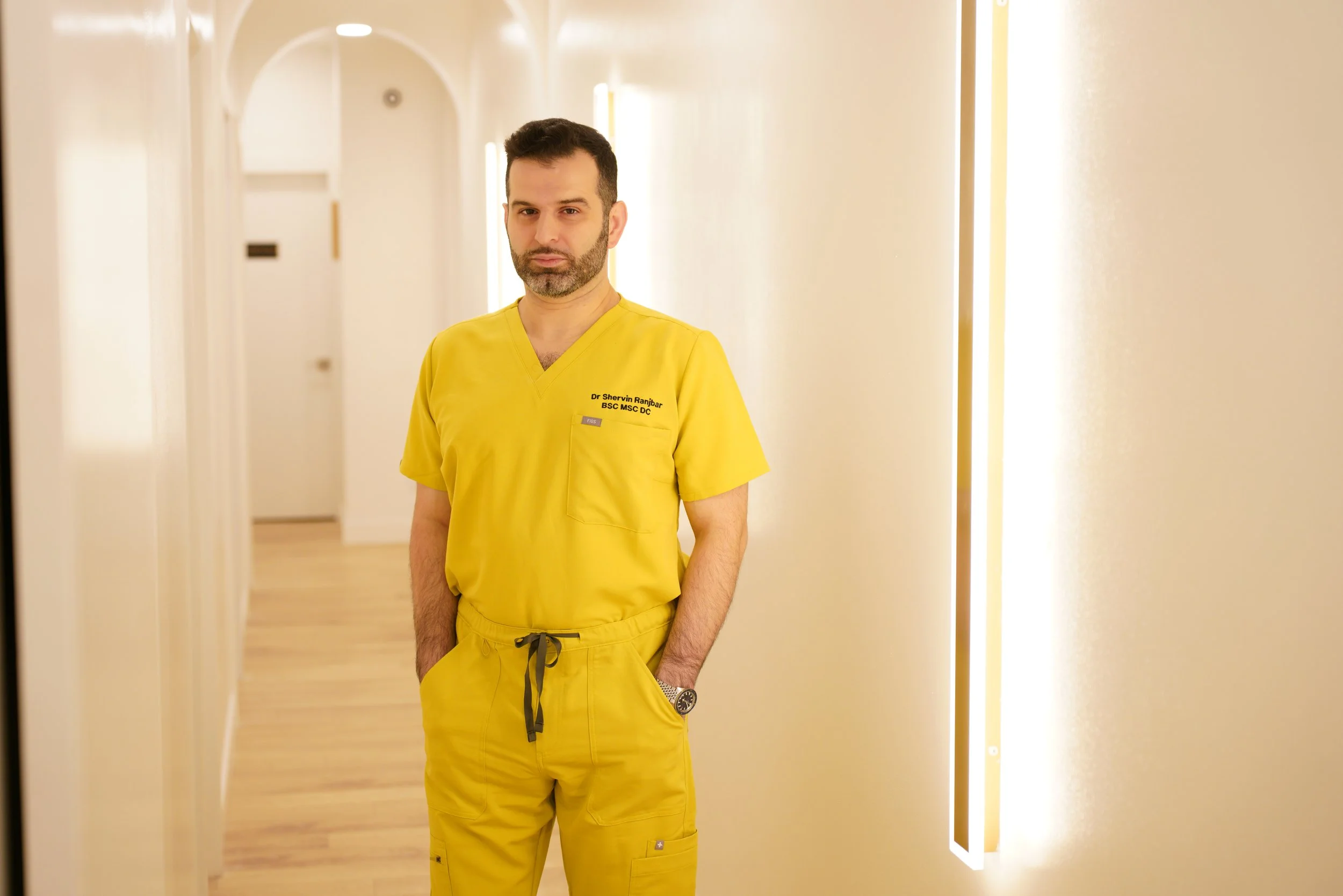 Dr Shervin Ranjbar in yellow scrubs standing with hands in pocket in hallway of near me therapy clinic cream walls wood coloured floor