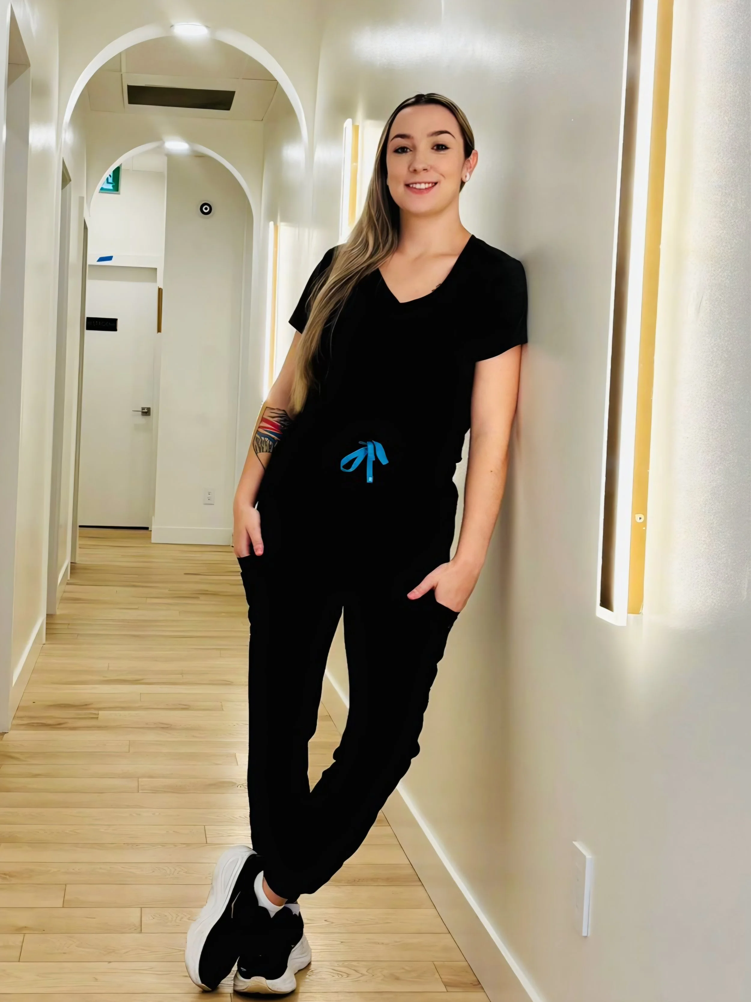 A young woman with long hair and tattoos on her left arm is smiling and leaning against a light-colored wall in a hallway with wooden floors, wearing a black shirt, black pants with a blue drawstring, and black sneakers with white soles.