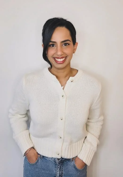 Young woman with dark hair, smiling, wearing a cream-colored cardigan and jeans, standing against a plain white wall.