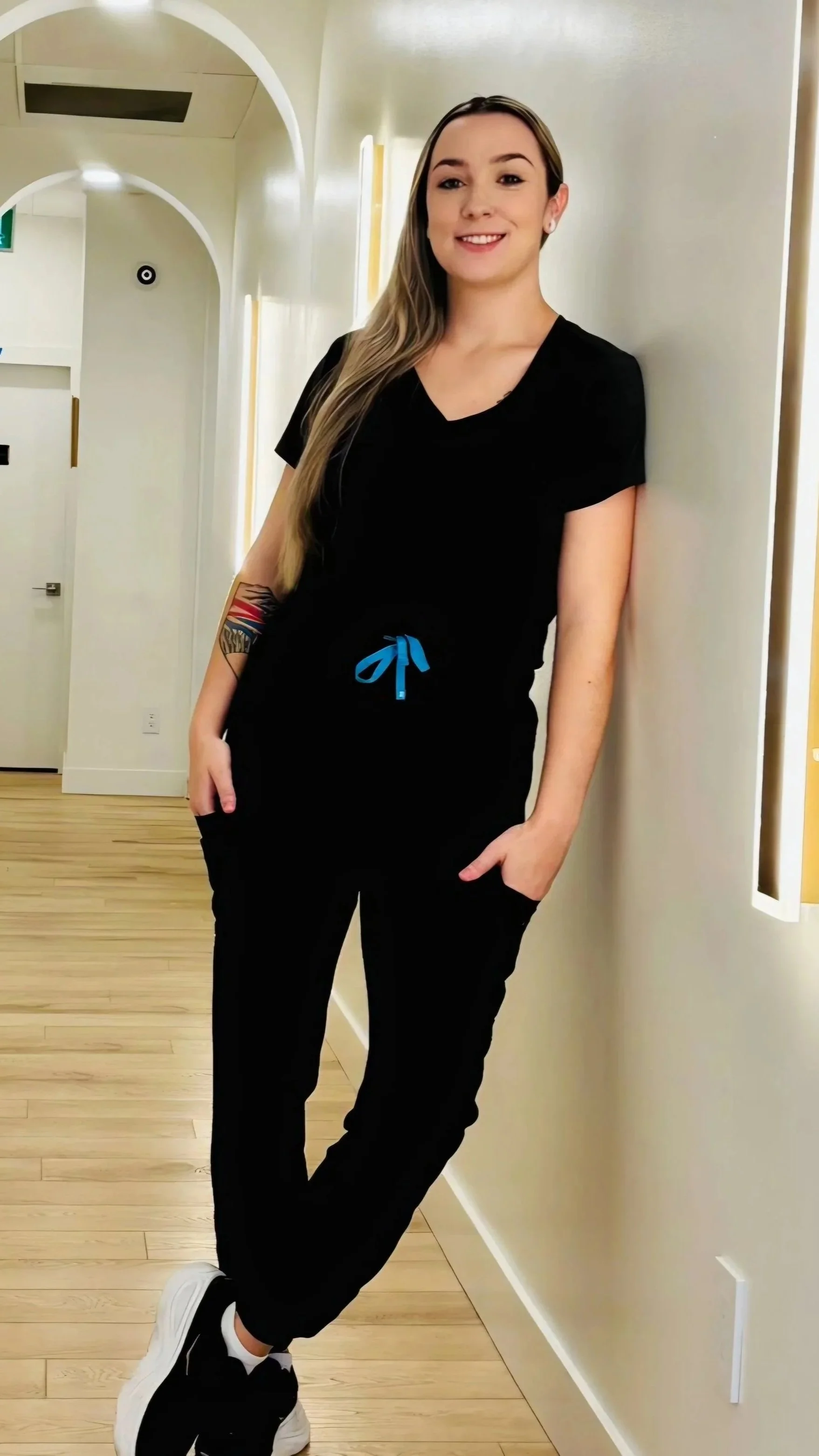 Mady the RMT at Near Me Therapy with long blonde hair wearing black scrubs with a blue drawstring, standing against a wall in a modern, brightly lit hallway with wood flooring.