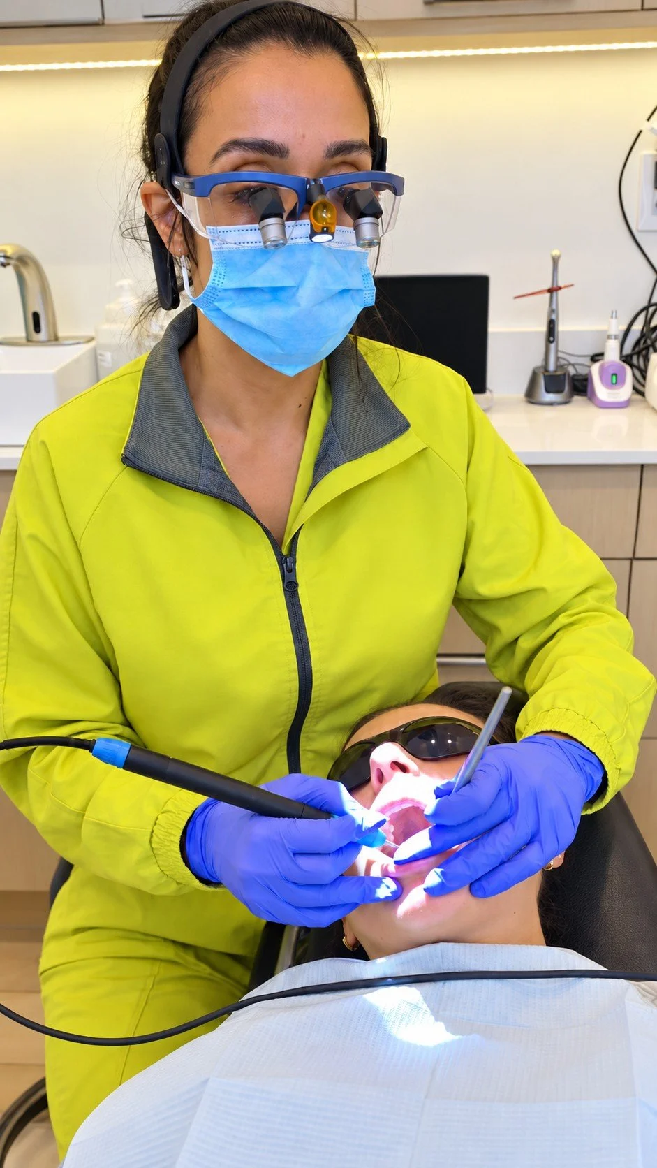 Kamelia bahrini dental hygienist performing dental work on a patient in an interdisciplinary office. The dental hygienist is wearing magnifying glasses, a mask, gloves, and a yellow dental coat, while the patient is lying back and an open mouth.