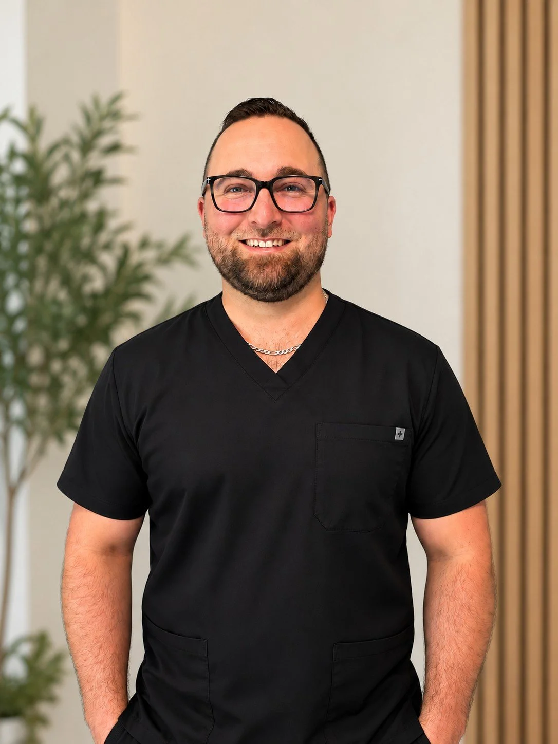 Tyson Popoff Acupuncturist in Langley City standing with Near me Therapy neutral woods in background and green plants wearing black scrubs and and a pair of glasses