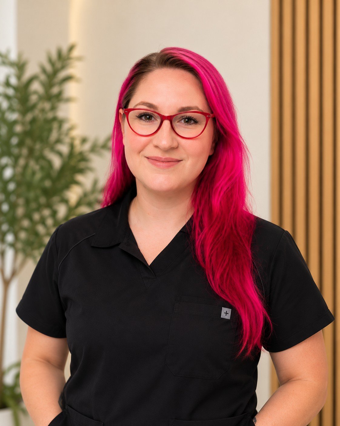 April Clarke standing with pink and black hair, red glasses and black scrubs with near me therapy plants and fluted wood background