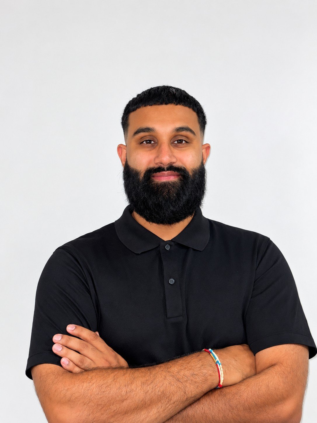 Portrait of a man with a beard and short hair, wearing a black polo shirt, standing with arms crossed, against a plain white background.