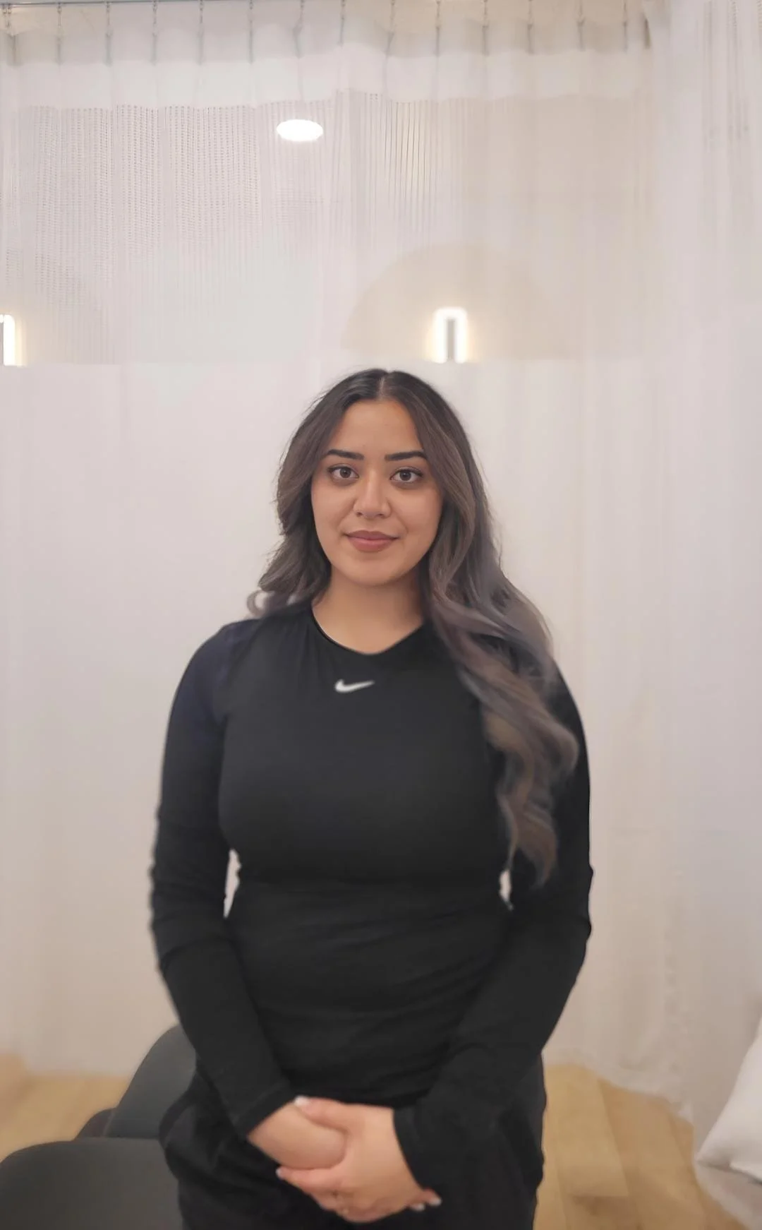 A young woman with long, wavy gray hair, wearing a black Nike long-sleeve top, sitting indoors in front of white curtains.