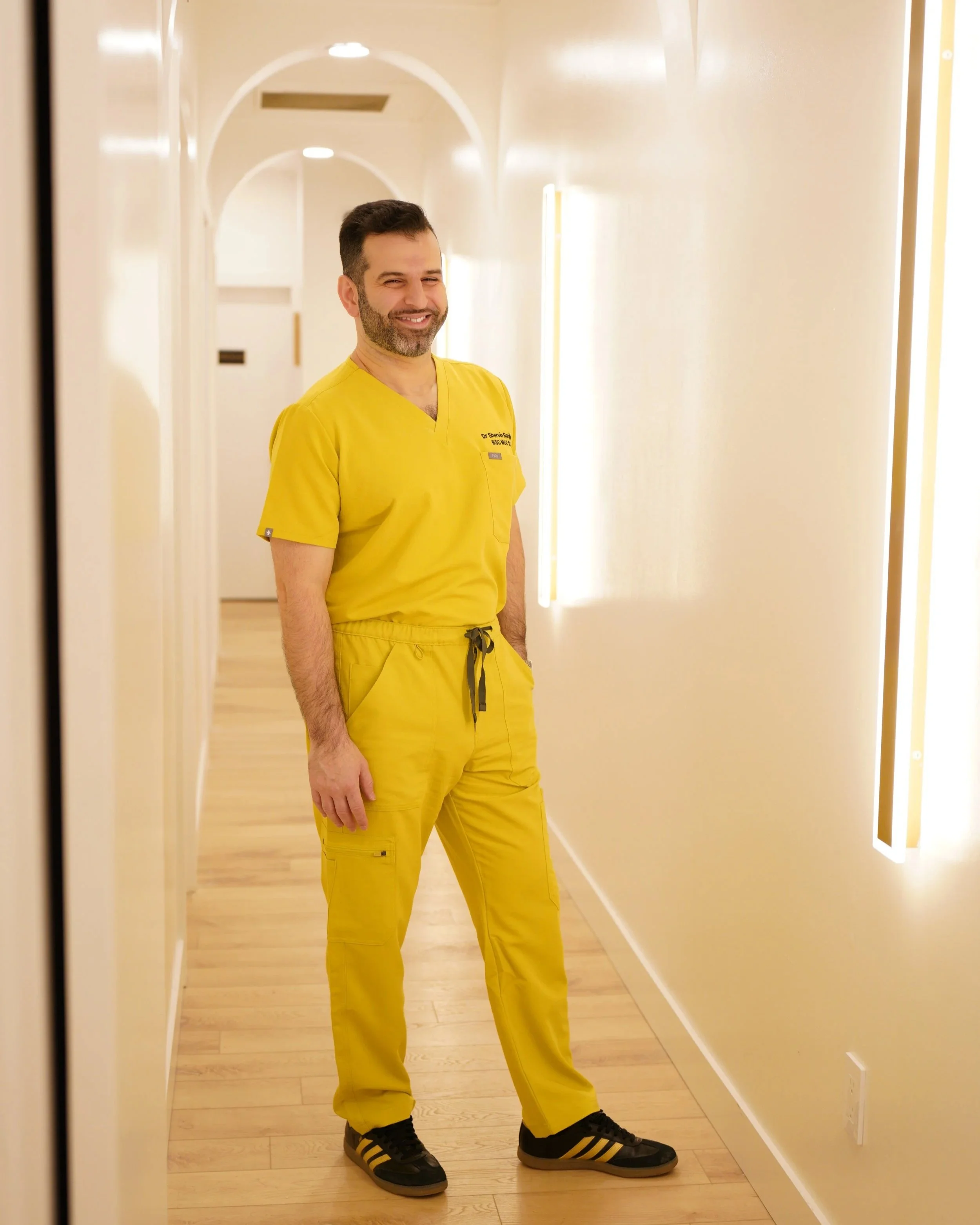Dr Shervin Ranjbar dressed in yellow medical scrubs standing in a brightly lit hallway with light-colored wood floors and white walls at Near Me Therapy Langley