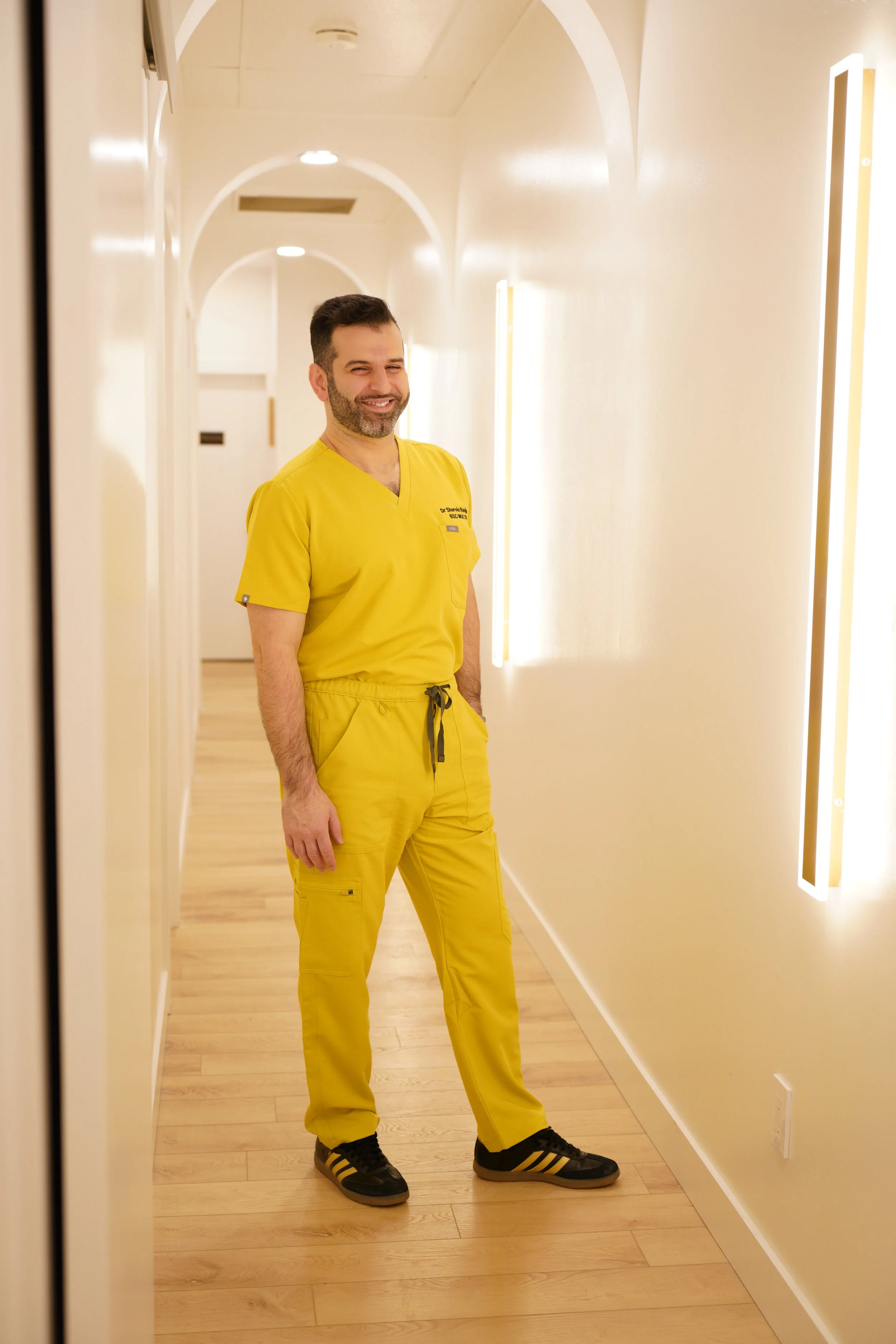 Dr Shervin Ranjbar, Chiropractor is standing in yellow scrubs in white hallway and wood coloured flooring with multiple sconces on the wall and arches above