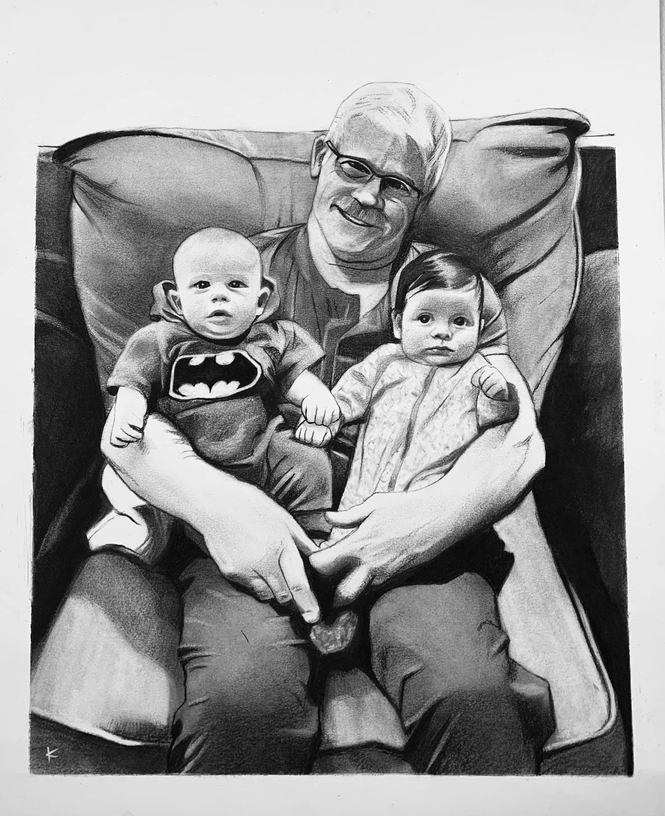 A photo of an older man with gray hair and glasses sitting on a sofa holding two babies, one in each arm. The baby on the left is wearing a Batman t-shirt, and the baby on the right is dressed in a patterned outfit.