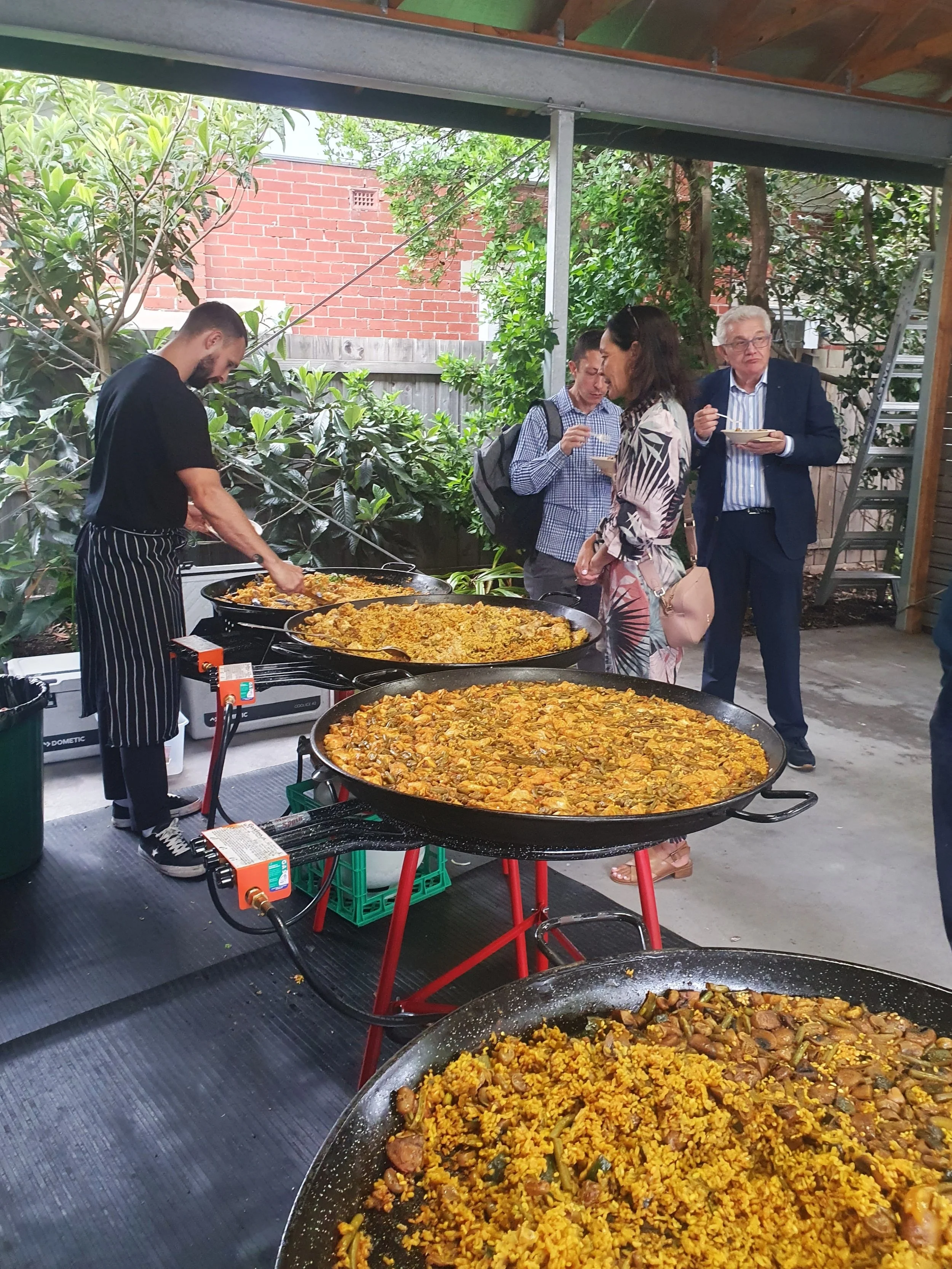 People at an outdoor food event, serving and eating large pans of paella, with a chef preparing the dishes.