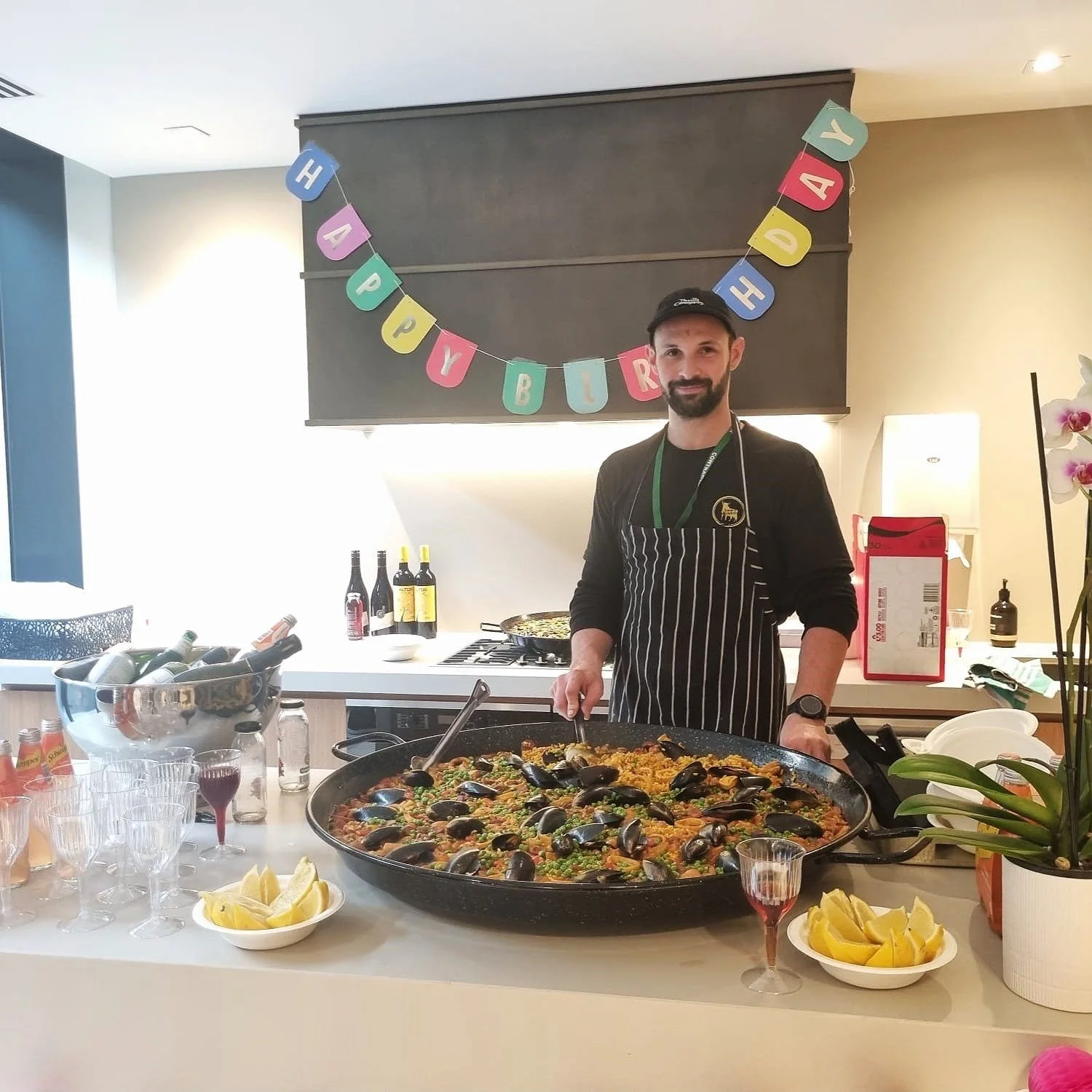 A man with a beard and a cap cooking seafood paella in a large pan at a celebration. Behind him is a birthday banner reading 'HAPPY BIRTHDAY'. The table has lemon wedges, glasses, wine bottles, and other party items.
