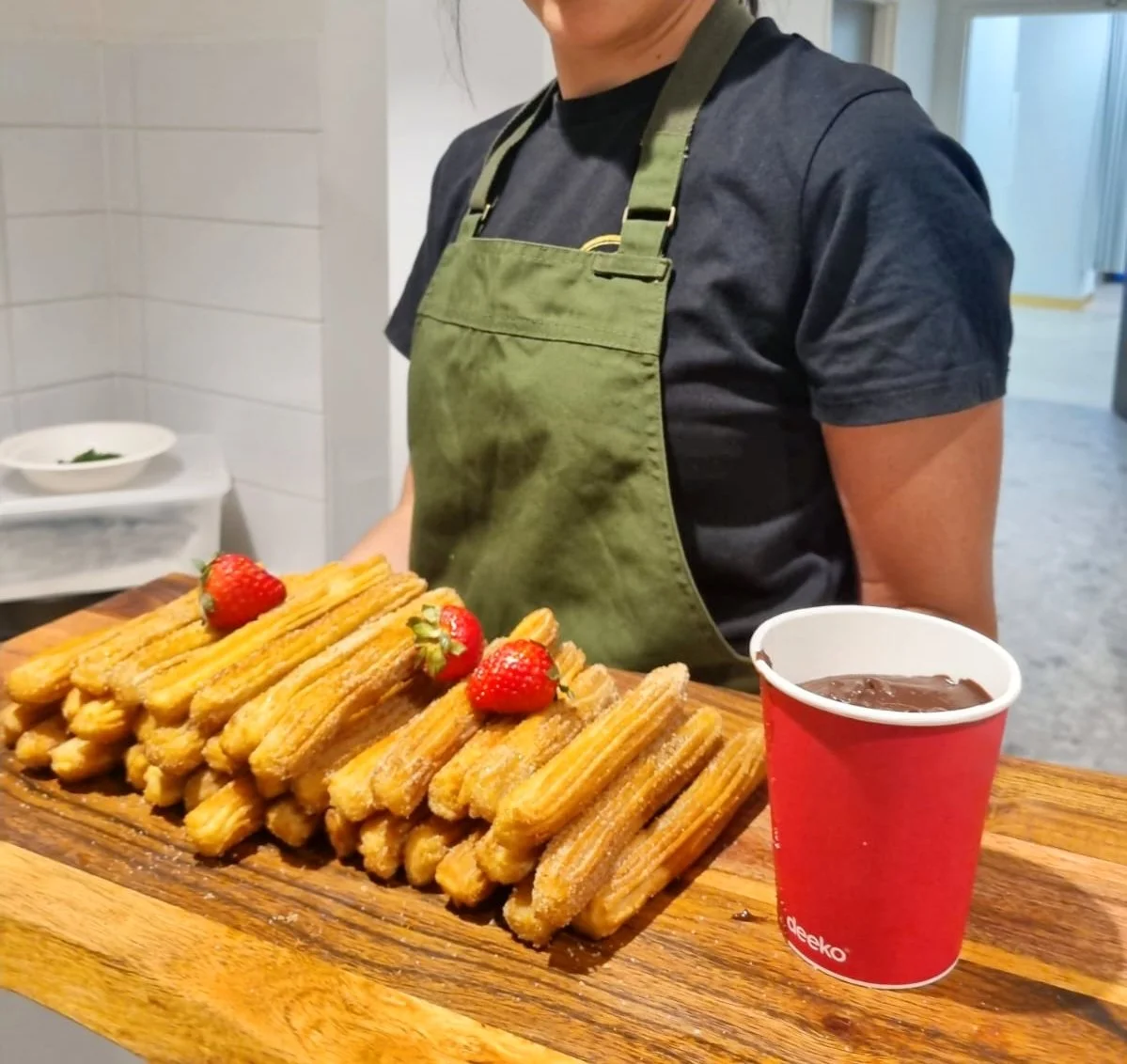 Person in black shirt and green apron stands behind a wooden table with a stack of churros topped with strawberries, a cup of hot chocolate, and a bowl in the background.