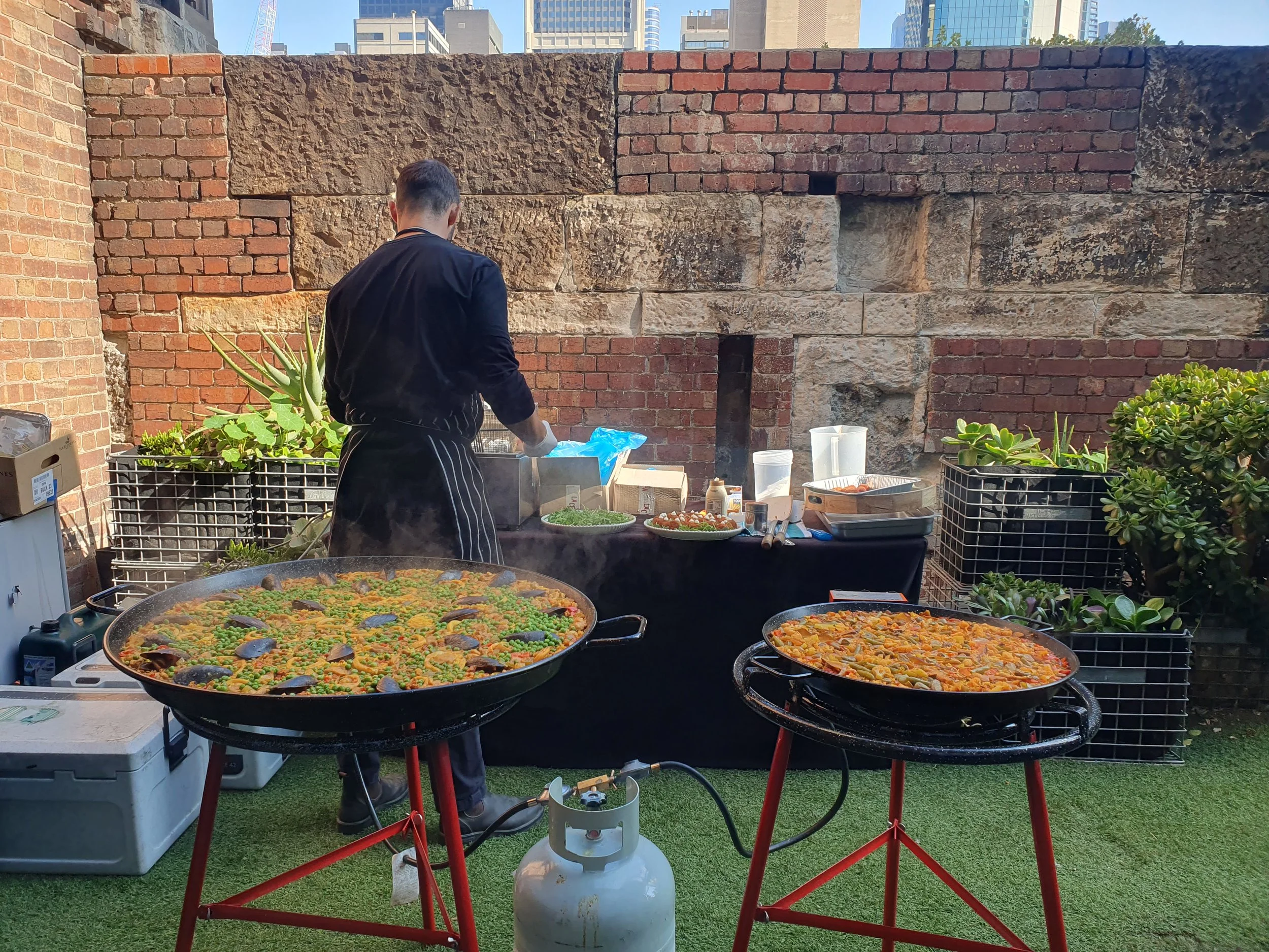 A chef cooking paella outdoors on two large pans, with various ingredients and cooking utensils on a black table. The scene is set against a brick wall with urban buildings in the background.