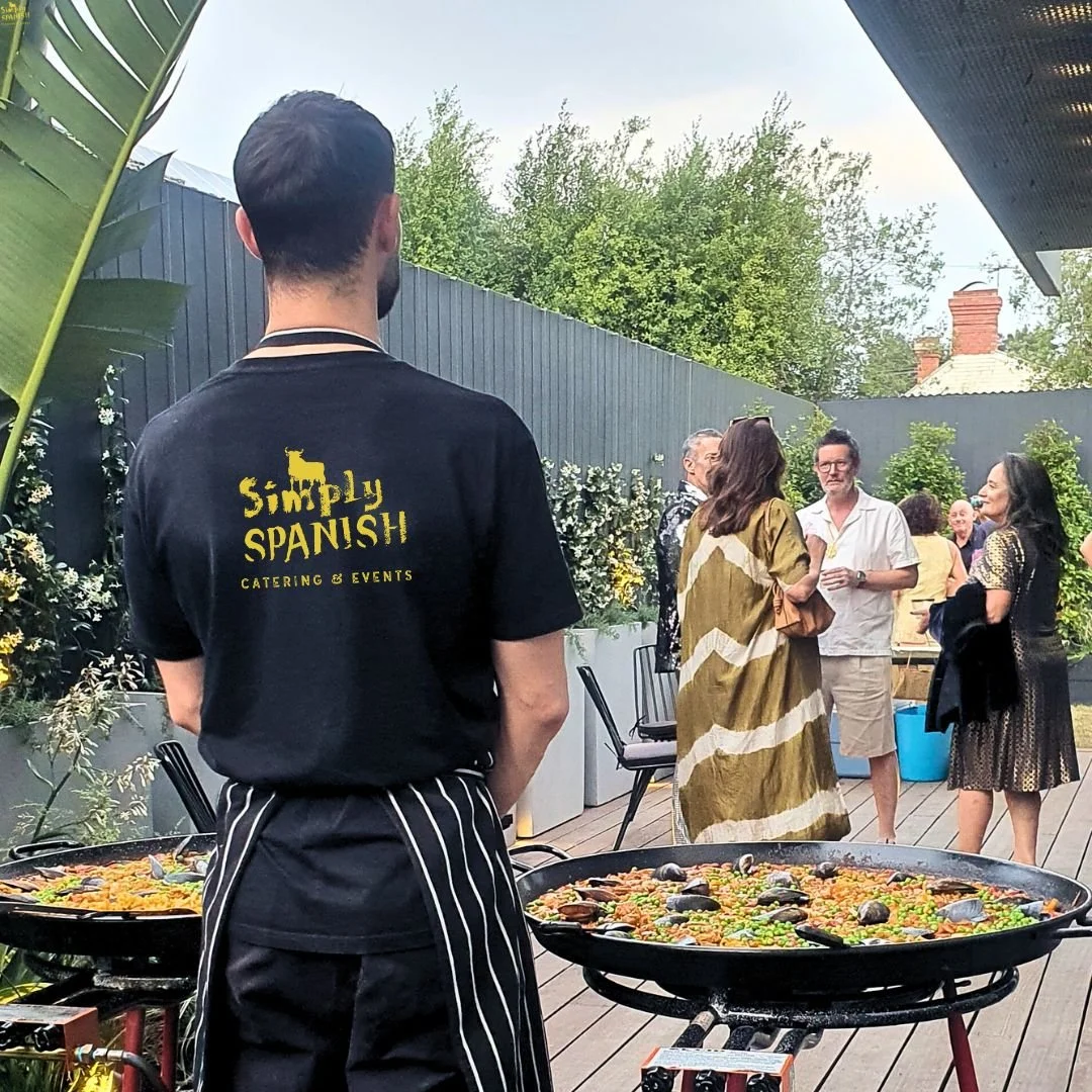 A man in a black Simply Spanish Catering & Events t-shirt and striped apron stands over a large paella pan on a wooden deck, with guests socializing in the background under a partly cloudy sky with greenery around.