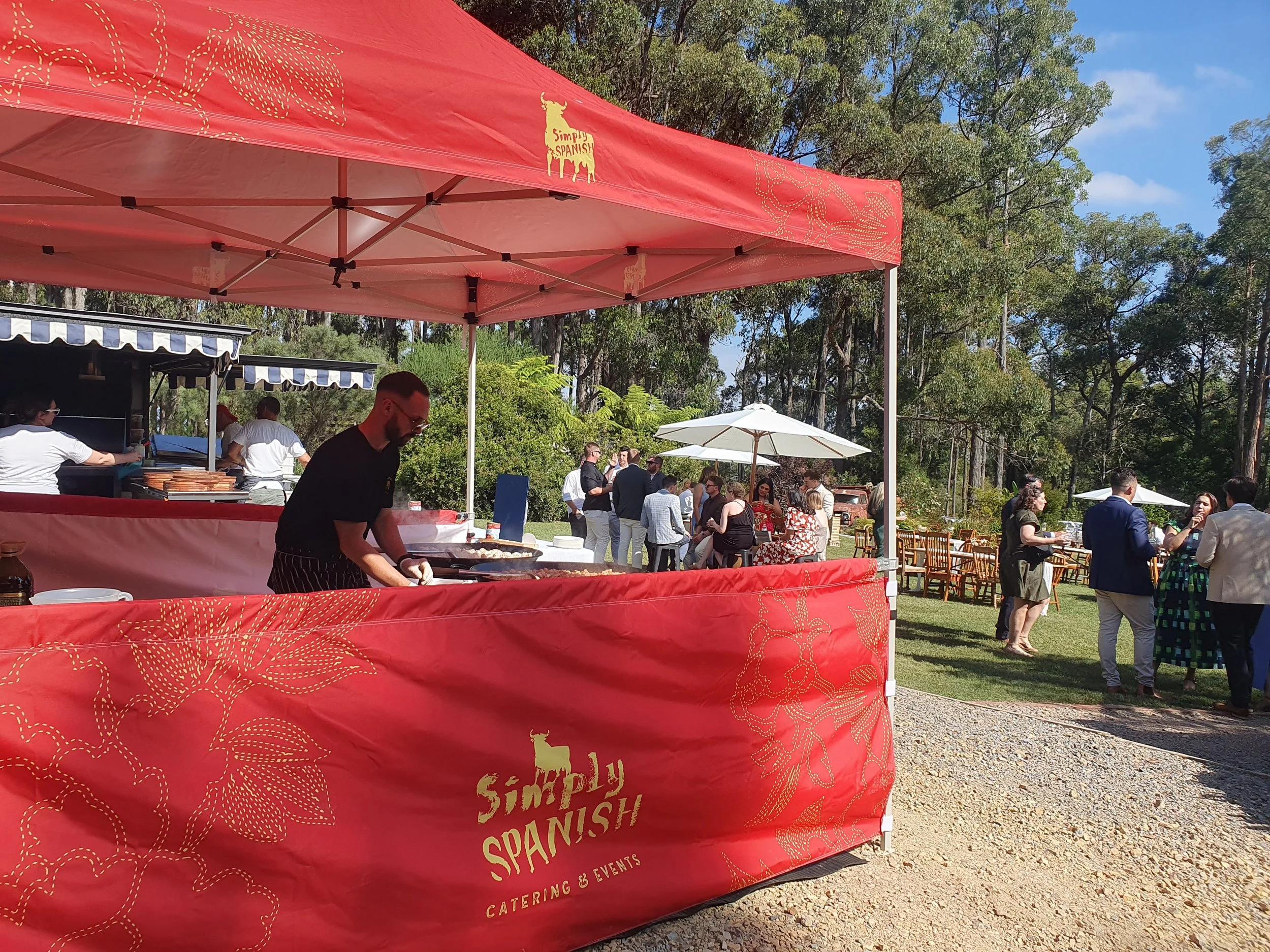 An outdoor catering event with a pink pavilion that has the words 'Simply Spanish Catering & Events' on it. Many people are gathered, some standing and chatting, others waiting at a food stand, with trees and blue sky in the background.