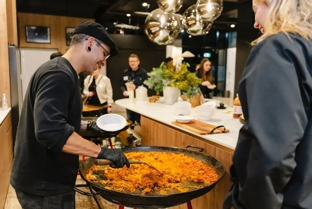 A chef serving paella to a customer at a restaurant bar, with people in the background and modern lighting decoration.