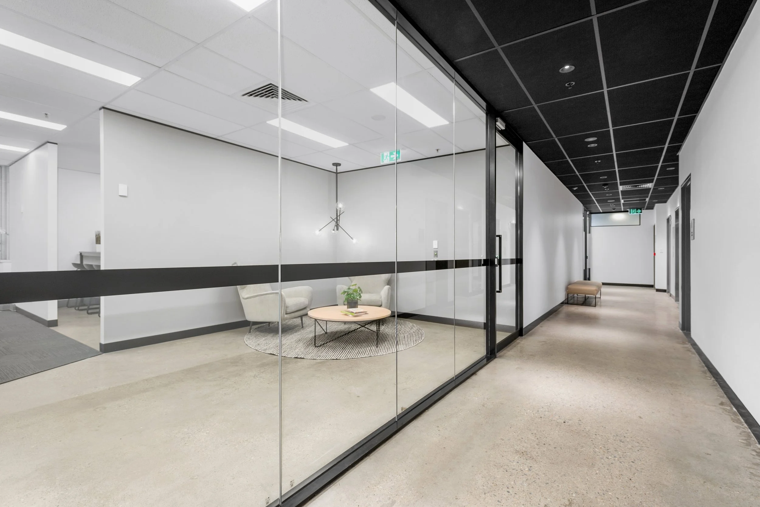 Modern office hallway with glass conference room on the left, white walls, black ceiling tiles, and beige flooring. There is seating along the right side of the hallway, and inside the conference room are two beige armchairs, a round coffee table wit