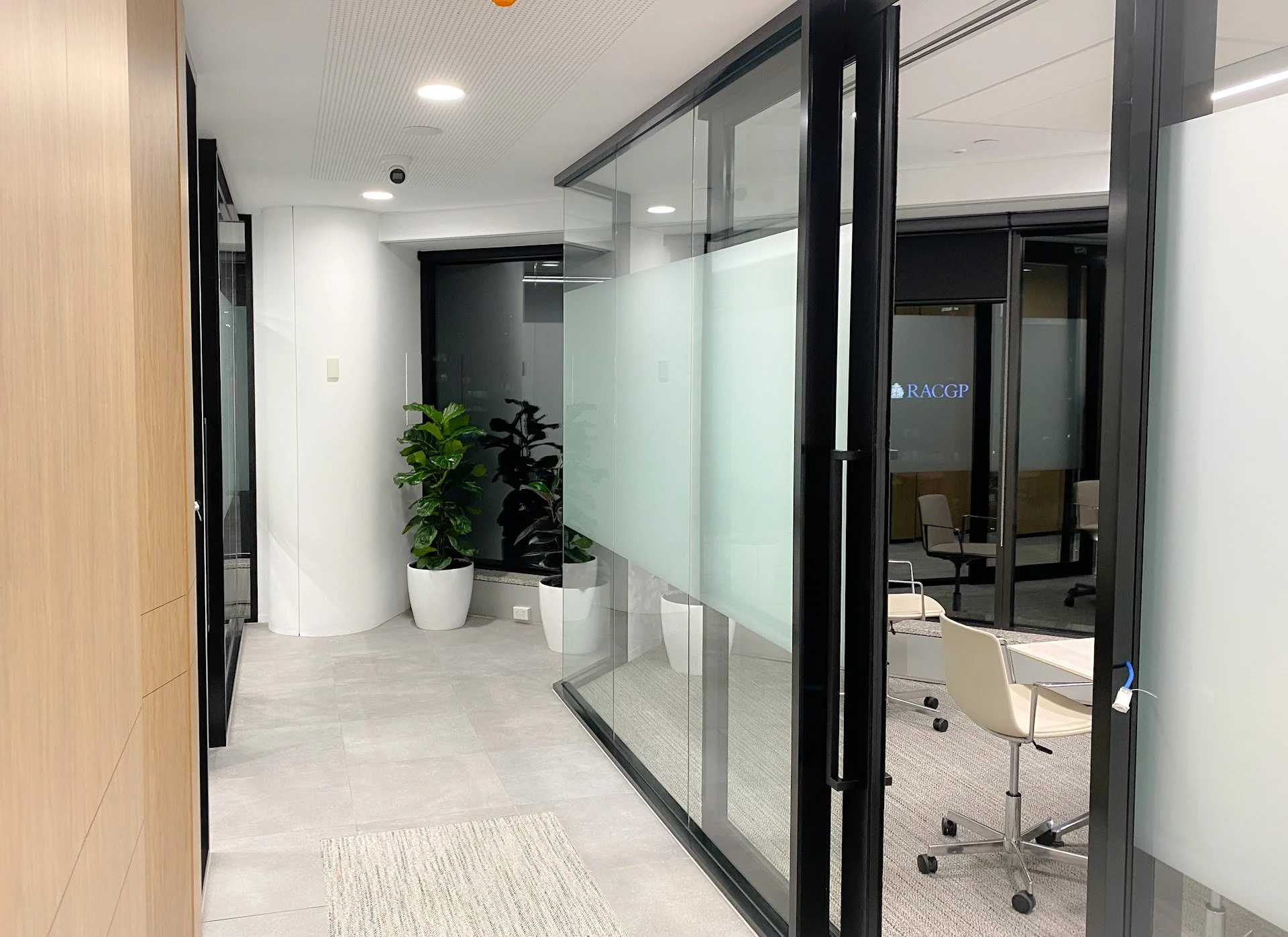 Modern office hallway with potted plants, glass walls, and conference room with chairs and a screen