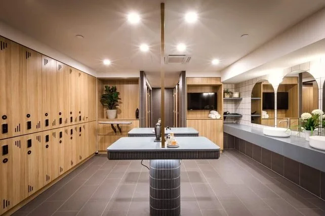 Modern locker room with wooden lockers, a shared table with two bottles, and a vanity area with sinks and mirrors.