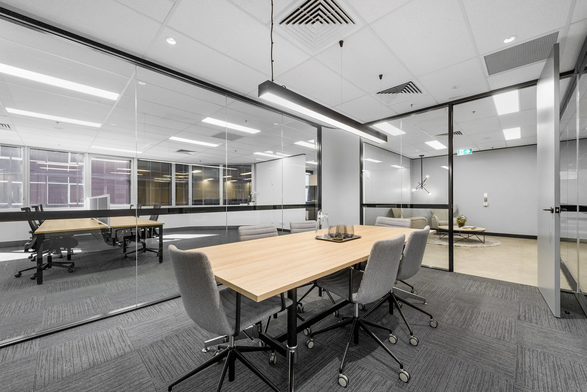 Interior view of a modern office conference room with a wooden table and gray chairs, separated from a lounge area by glass walls, featuring a sofa, chairs, and a coffee table.