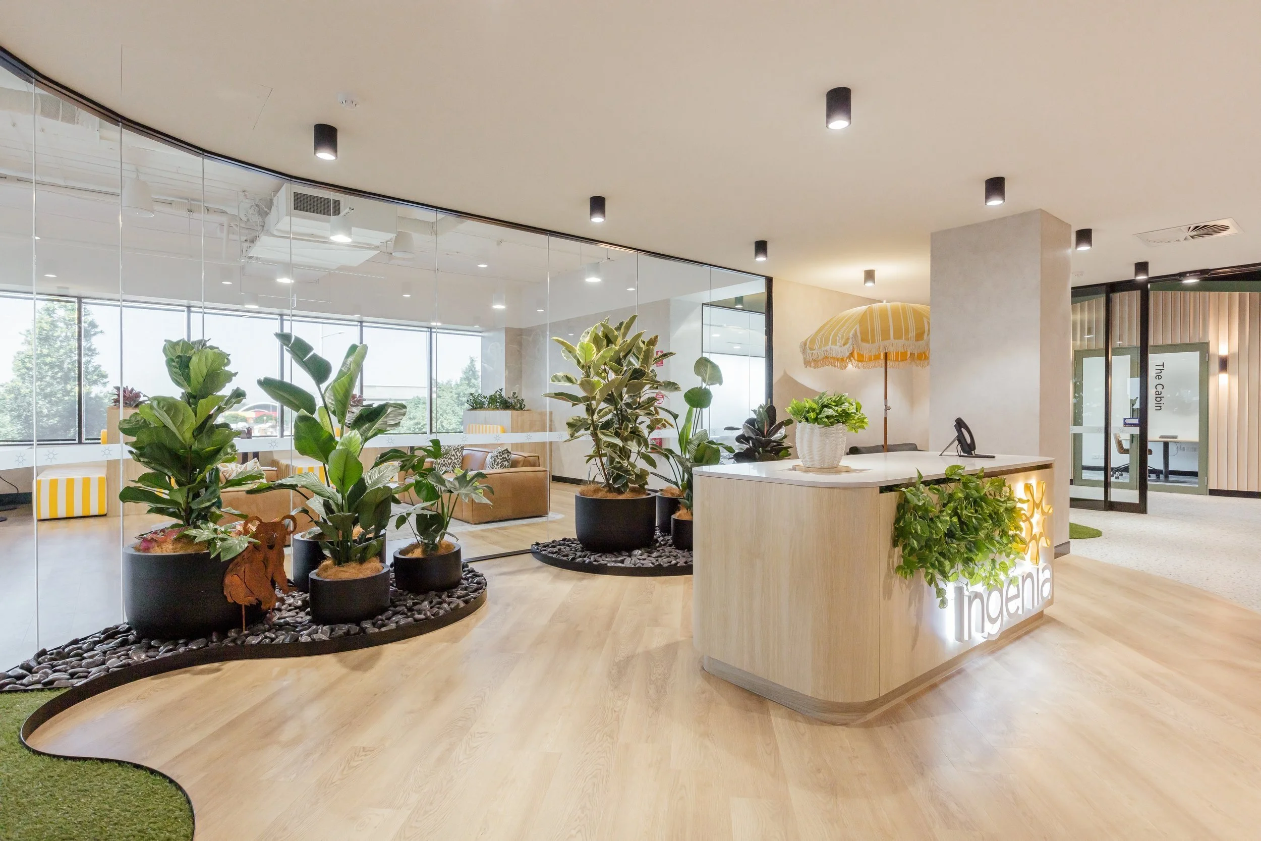 Modern office reception area with a curved wooden desk, illuminated logo, potted plants, large glass windows, and seating area with yellow striped furniture.
