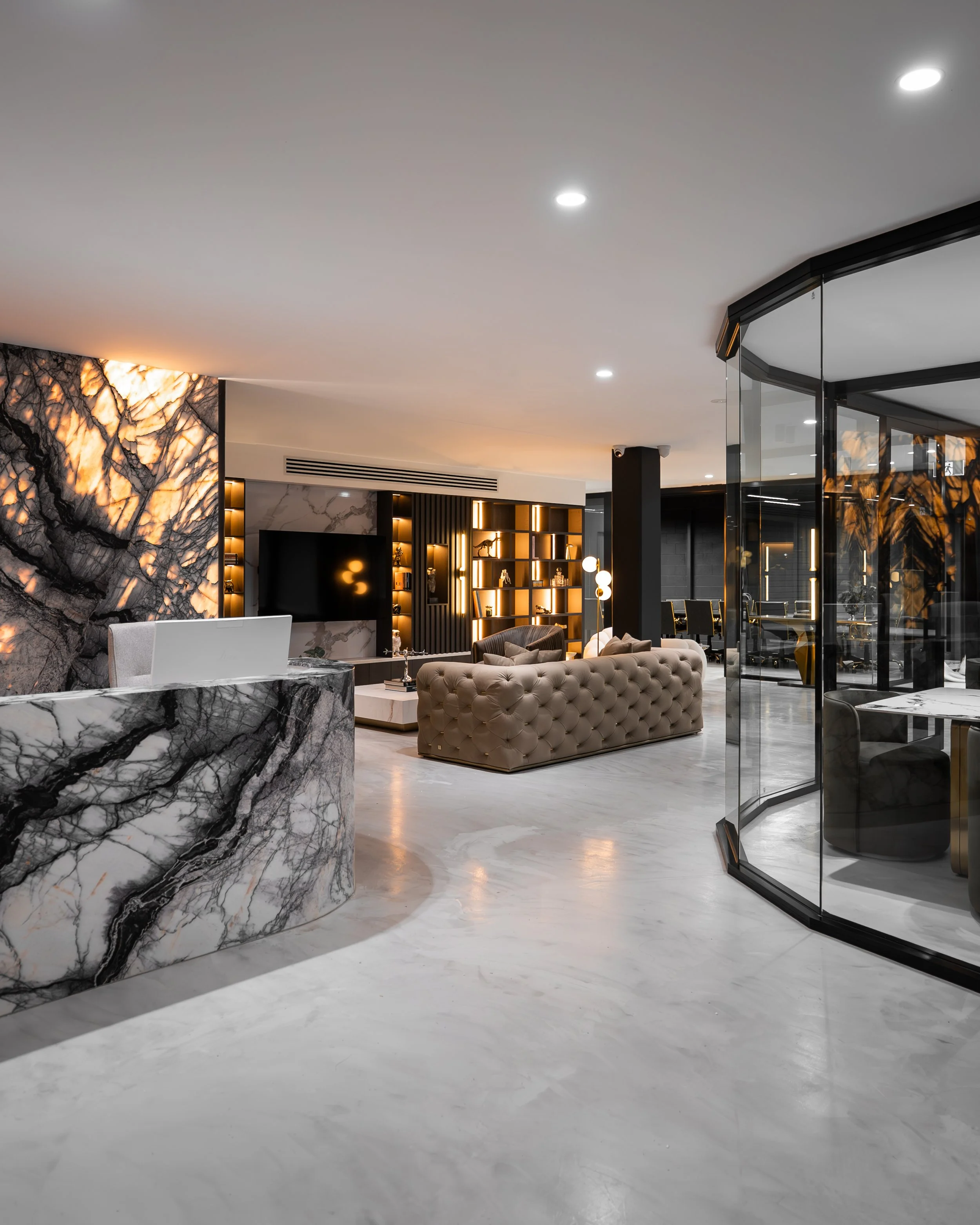 Modern hotel lobby with marble reception desk, tufted beige sofa, and glass-walled meeting rooms, featuring black and gold accents and warm lighting.