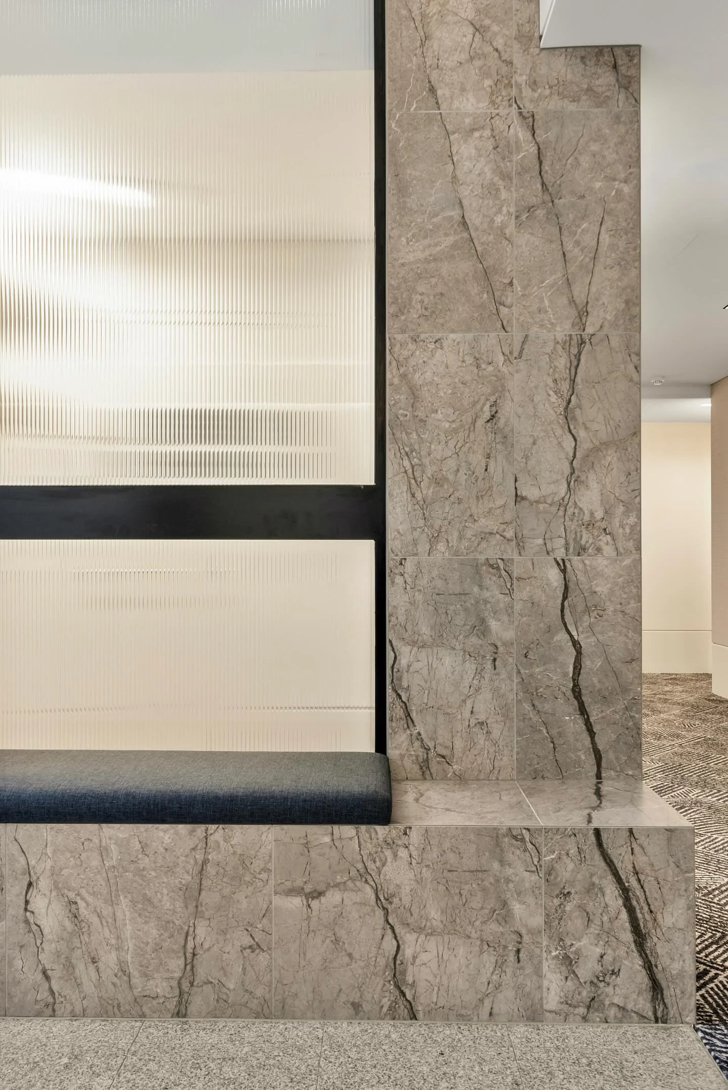 Interior view of a modern building with beige marble wall tiles, a frosted glass panel, and a cushioned bench with dark fabric.