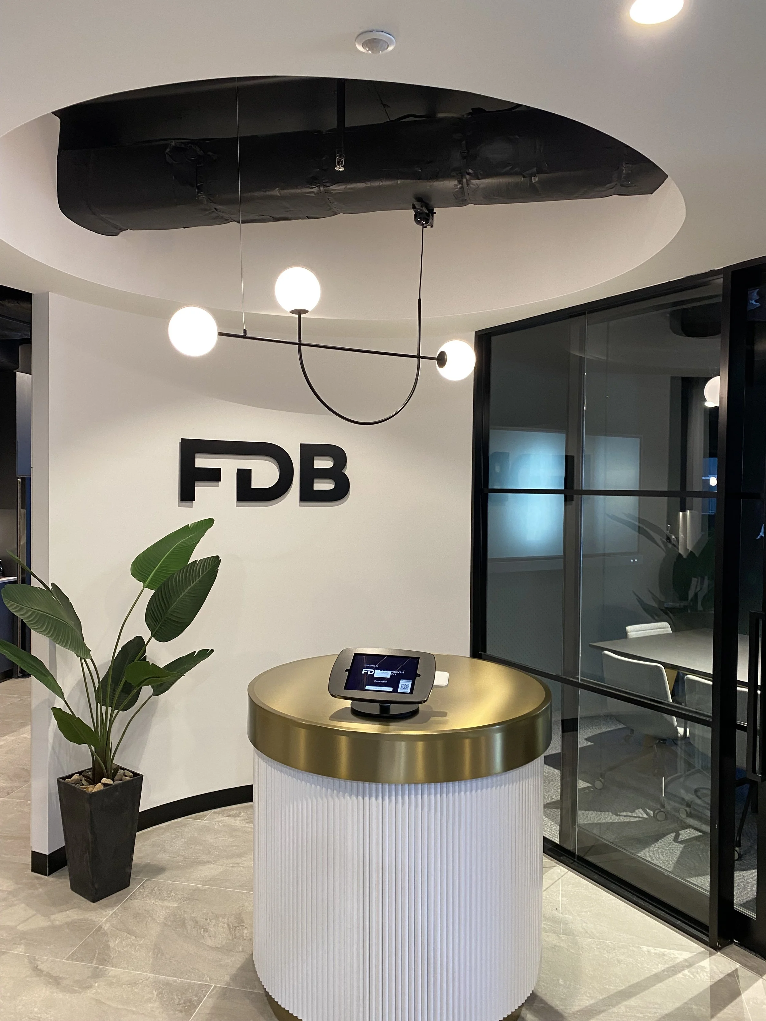 Interior of a modern office reception area with a white curved desk with gold top, digital check-in kiosk, large potted plant, and black wall with the letters FDB. Modern lighting fixtures hang from the ceiling.