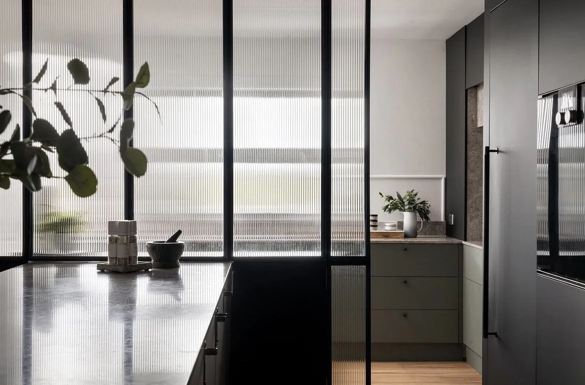 Modern kitchen with black cabinets, a grey countertop, and decorative plants, separated by a glass partition with vertical ridged texture.