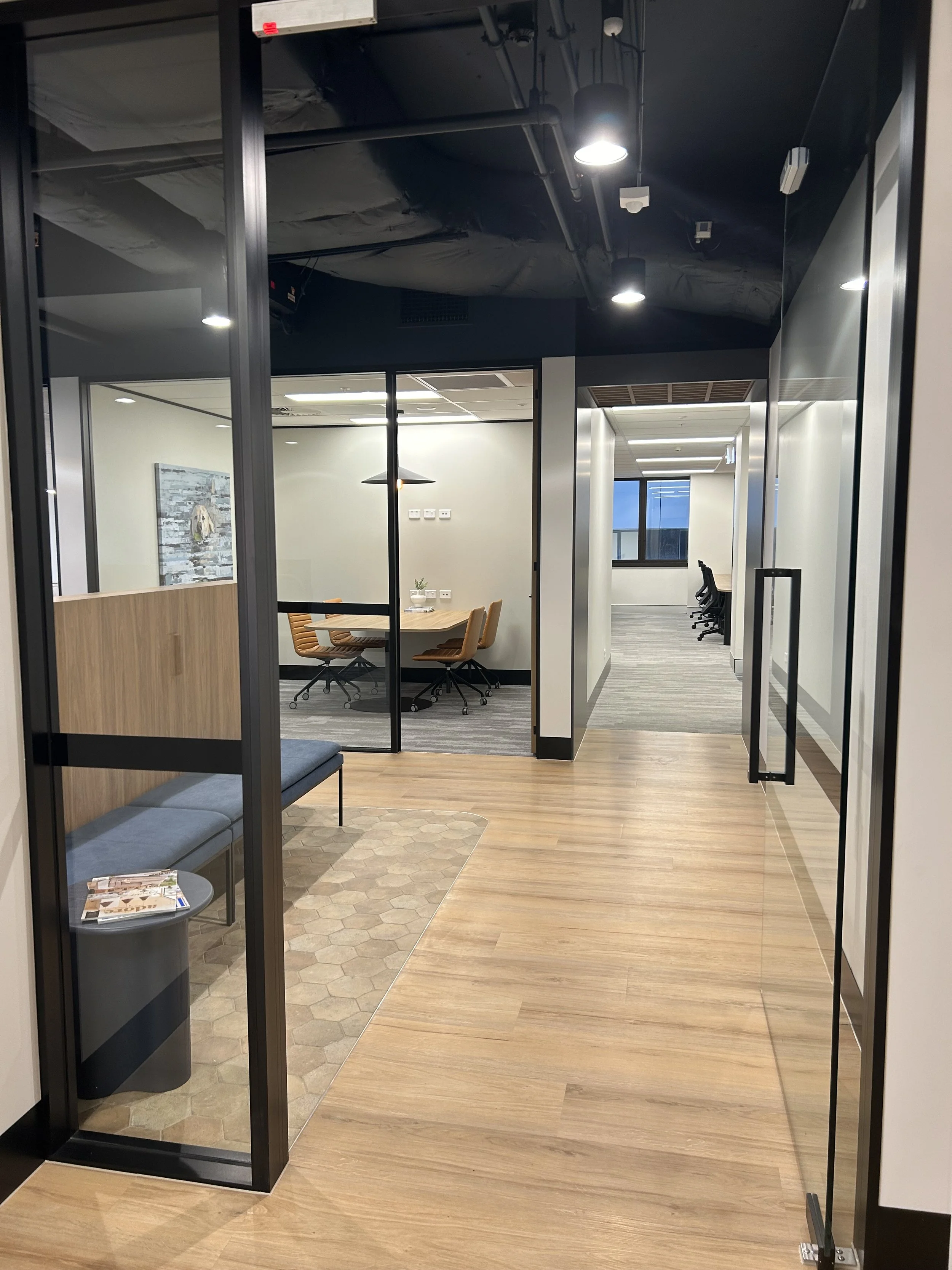 Modern office corridor with glass-walled conference rooms, wooden flooring, and contemporary furniture.