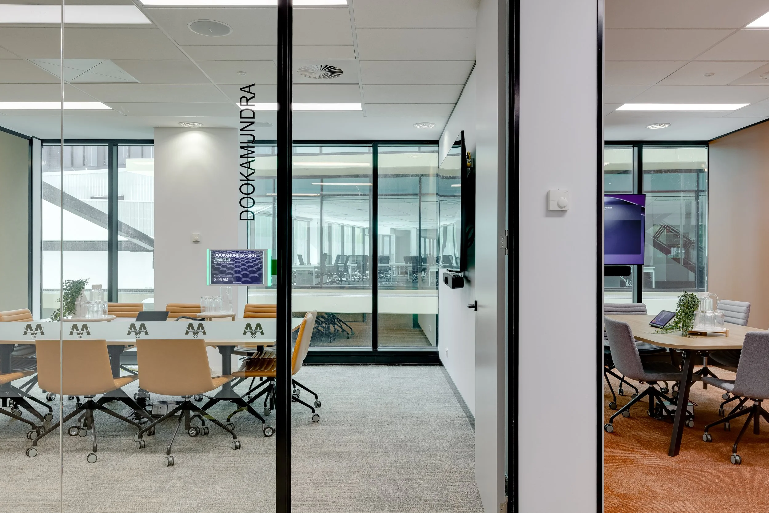 Modern conference rooms separated by glass walls with tables, chairs, laptops, and digital displays in an office setting.