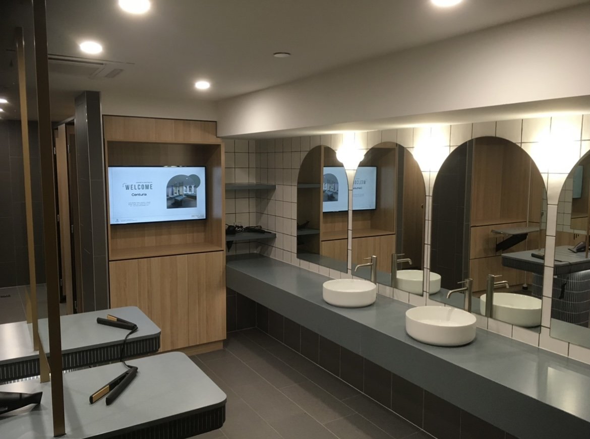 Public restroom with three white vessel sinks, mirrors, wooden panel accents, tiled walls, and a flat screen display with a welcome message.
