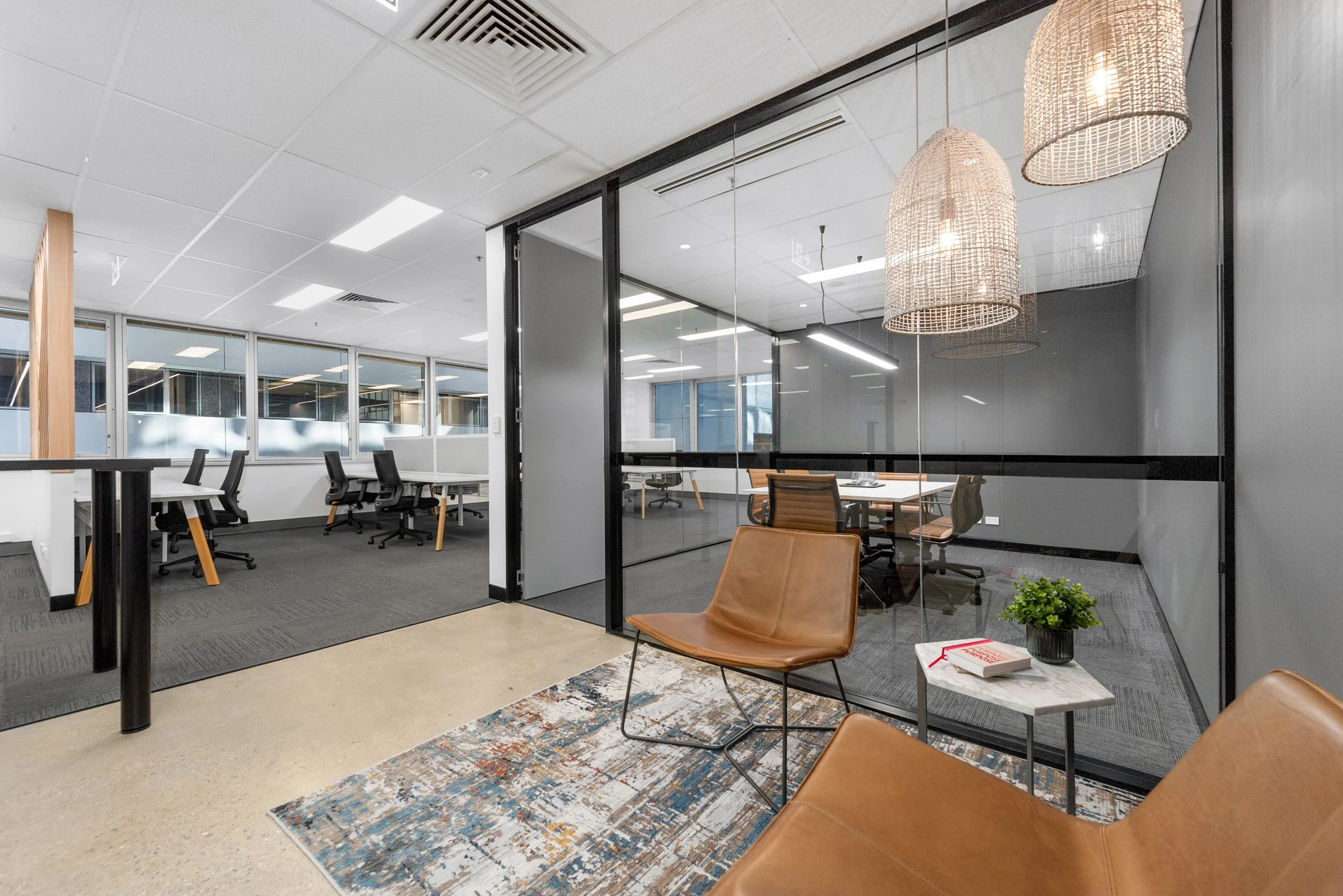 Modern office space with meeting rooms and workstations, leather chairs, a small side table with a book and a potted plant, pendant lights, large windows, and a colorful rug.