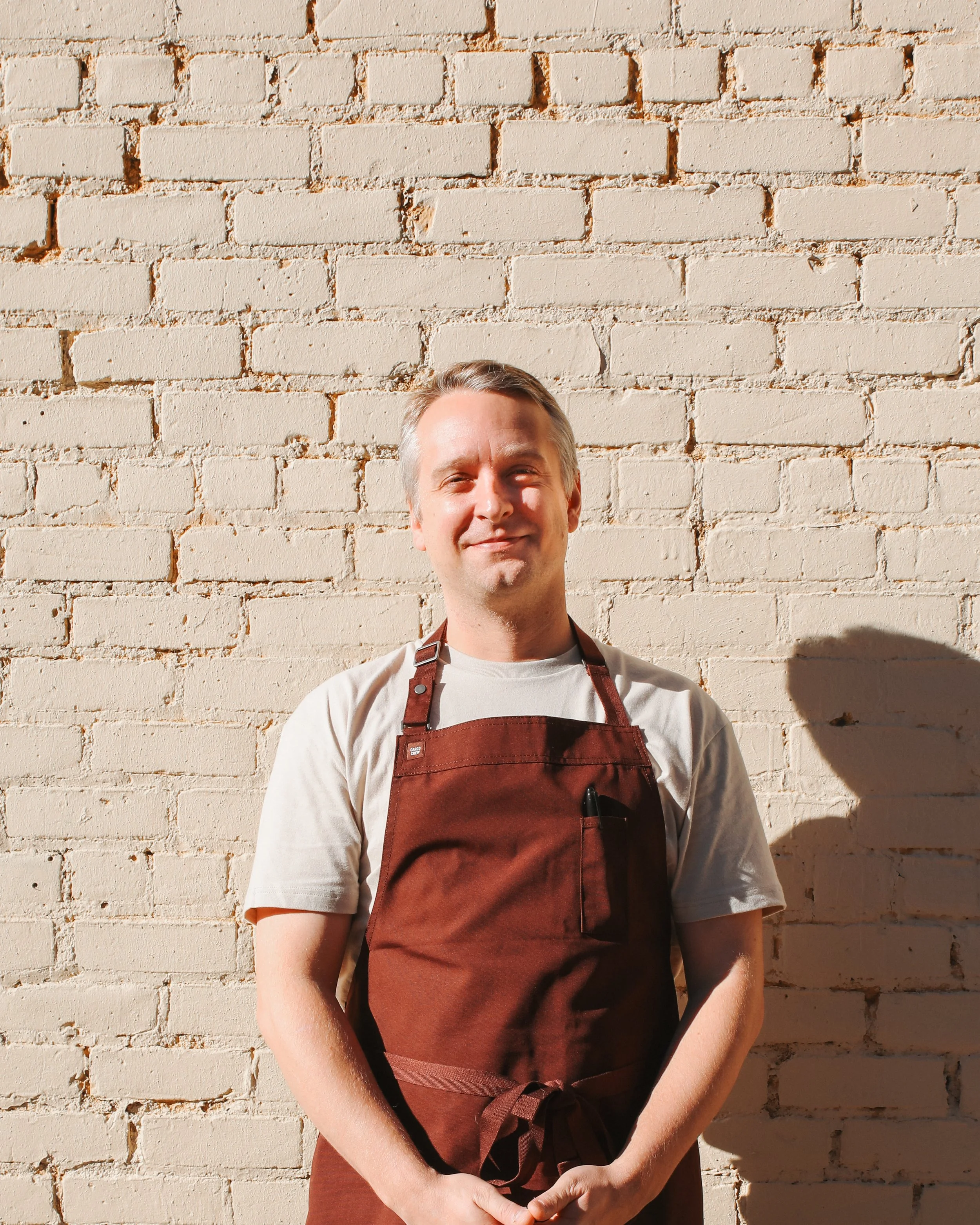 Chef Bjorn, from the Mexico en la Piel event, wearing a brown apron standing against a sunlit white brick wall