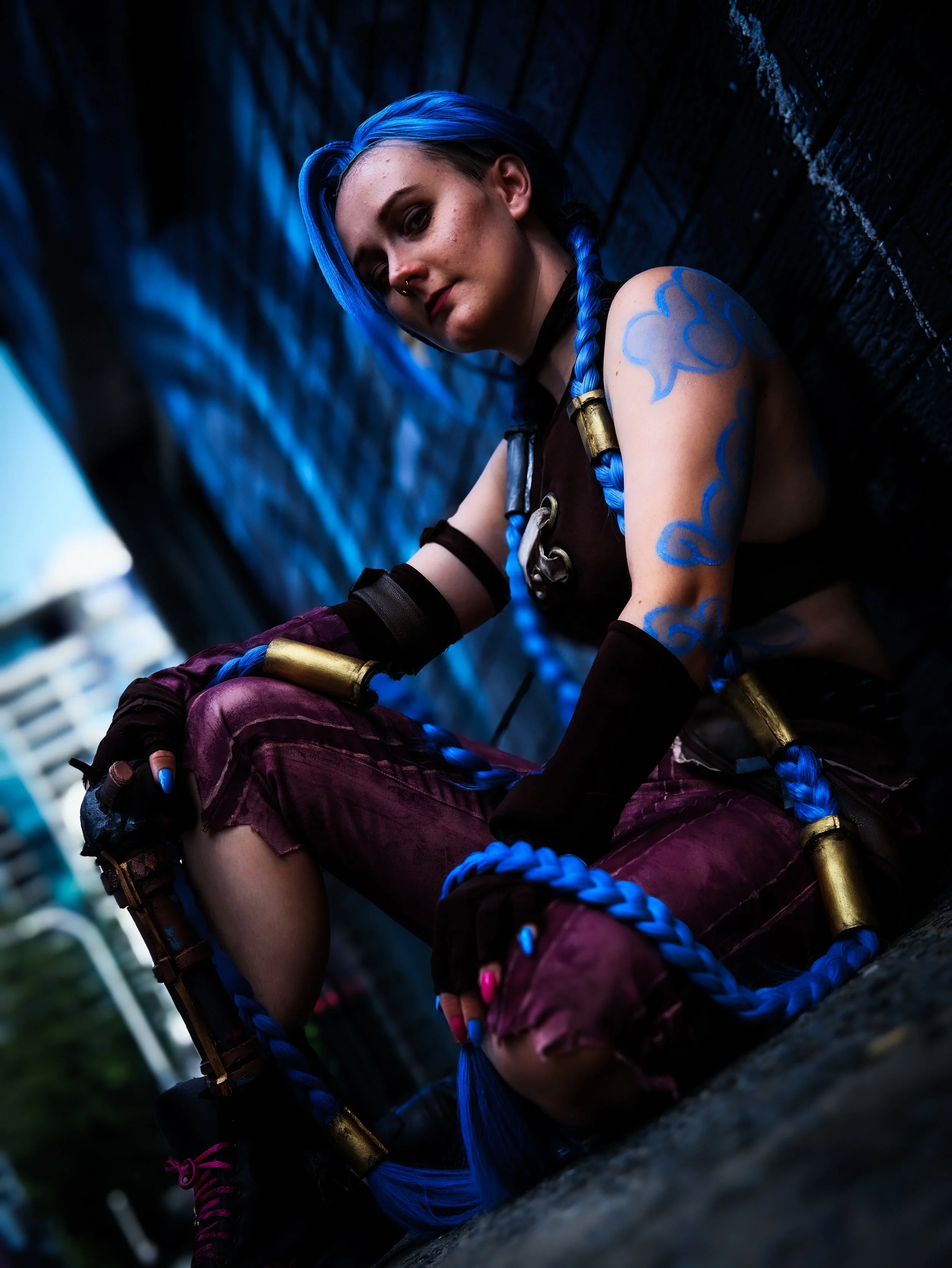 A character with blue braided hair and blue cloud tattoos on her arm sitting against a painted black brick wall in an urban setting.